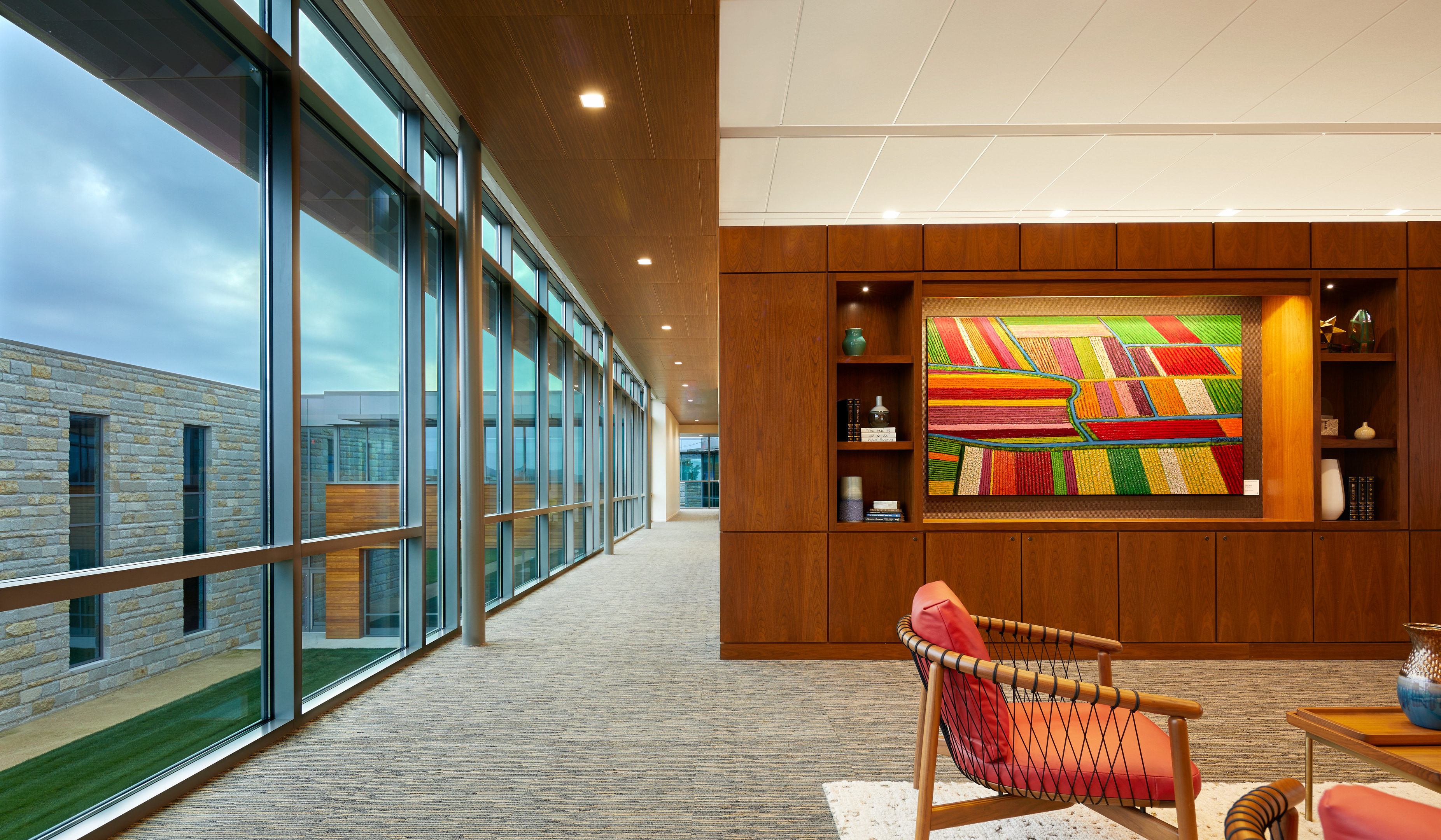 A modern hallway with large windows on one side and a wooden wall adorned with a colorful abstract painting on the other. The hallway, reminiscent of a research center, is furnished with chairs and decor items.