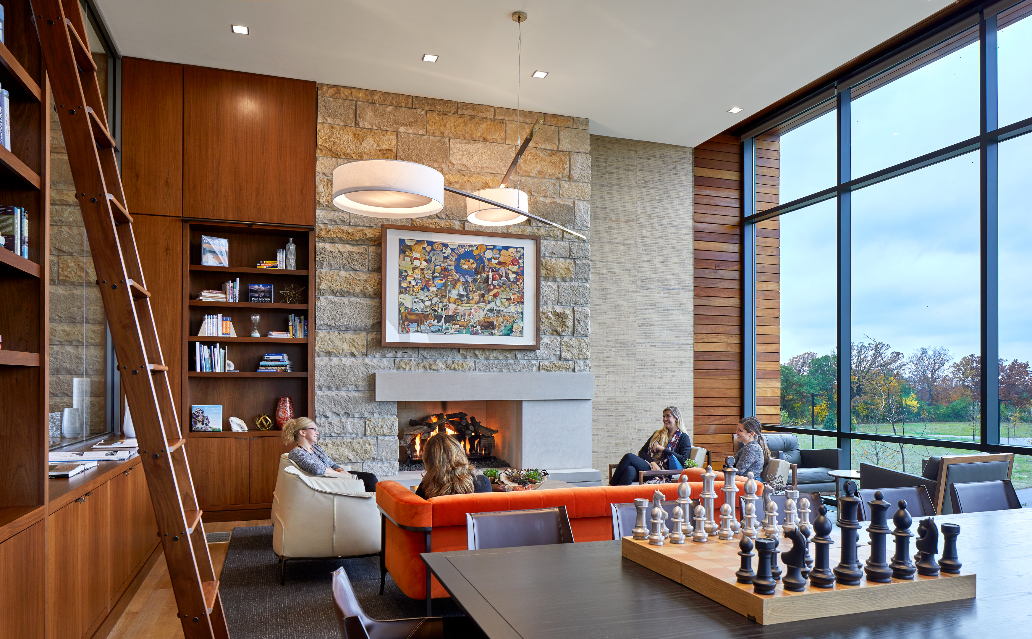 A modern living room with a high ceiling, a large bookcase with a ladder, a fireplace, and a large window offering a view of the outdoors. Several people are seated on an orange couch and chairs, making the space feel like an inviting home office.