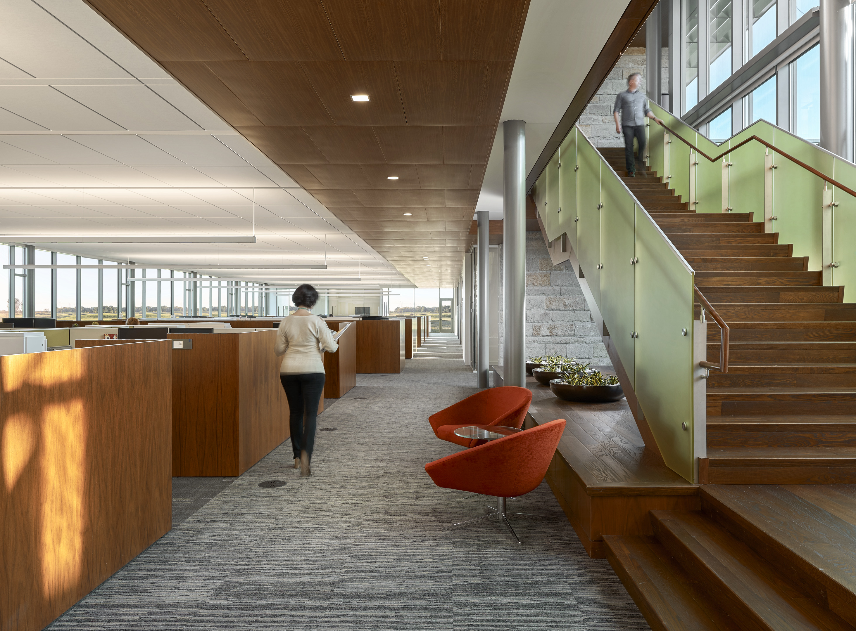 A spacious, modern office space with open cubicles on the left, two red chairs in the center, and a person descending a large wooden staircase on the right. Another person walks towards the cubicles, creating an atmosphere that blends the comfort of a Home Office with professional efficiency.