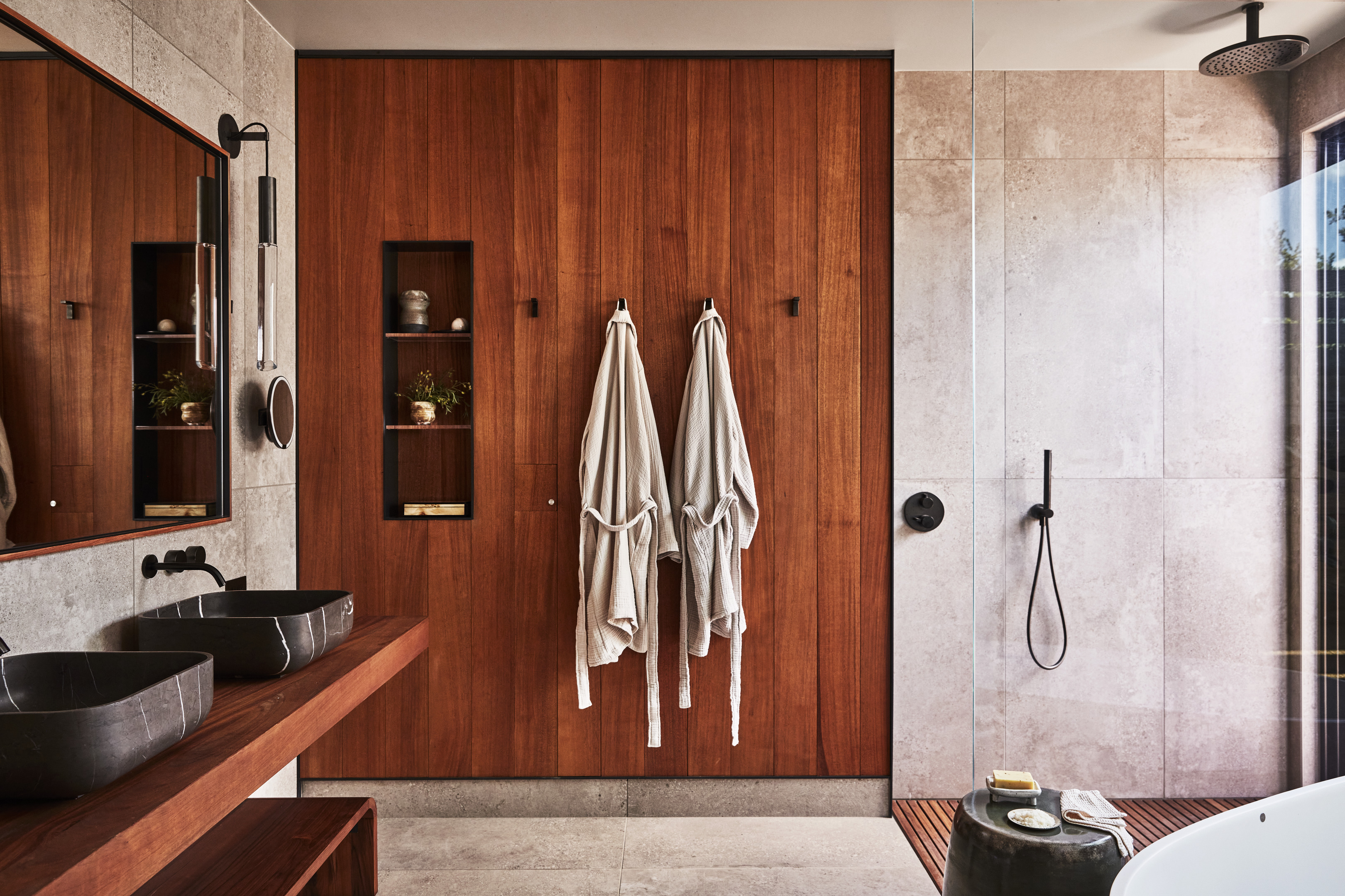 A modern bathroom at Stanly Ranch, part of the Auberge Resorts Collection, features wooden cabinetry, two black sinks, wall-mounted mirrors, robes on hooks, a shower with a rain showerhead, and a bathtub with a small stool beside it.