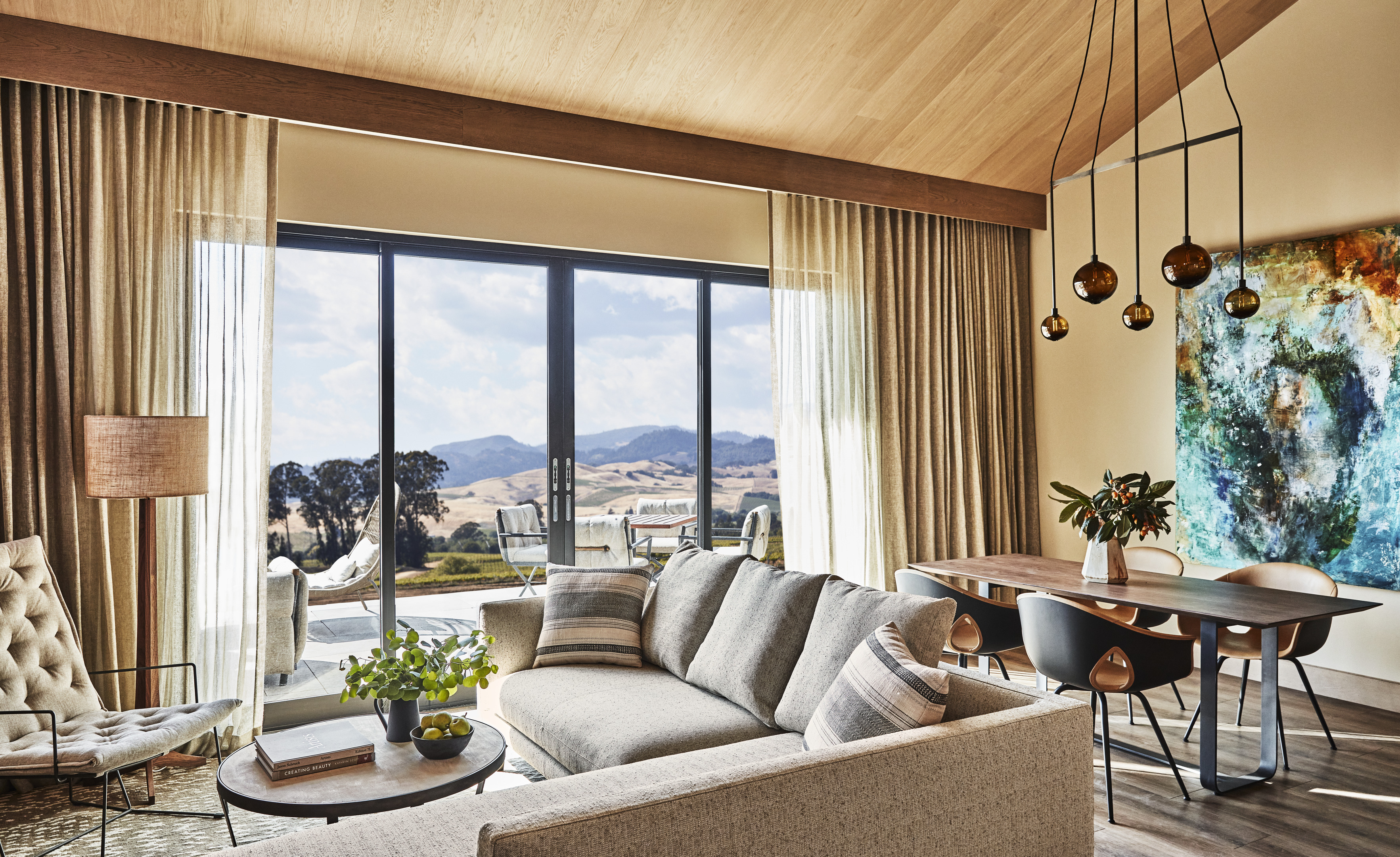 Modern living room with large windows, mountain views, neutral sofas, a wooden dining table, abstract wall art, pendant lights, and natural decor elements reminiscent of luxury resorts like Stanly Ranch by Auberge Resorts.