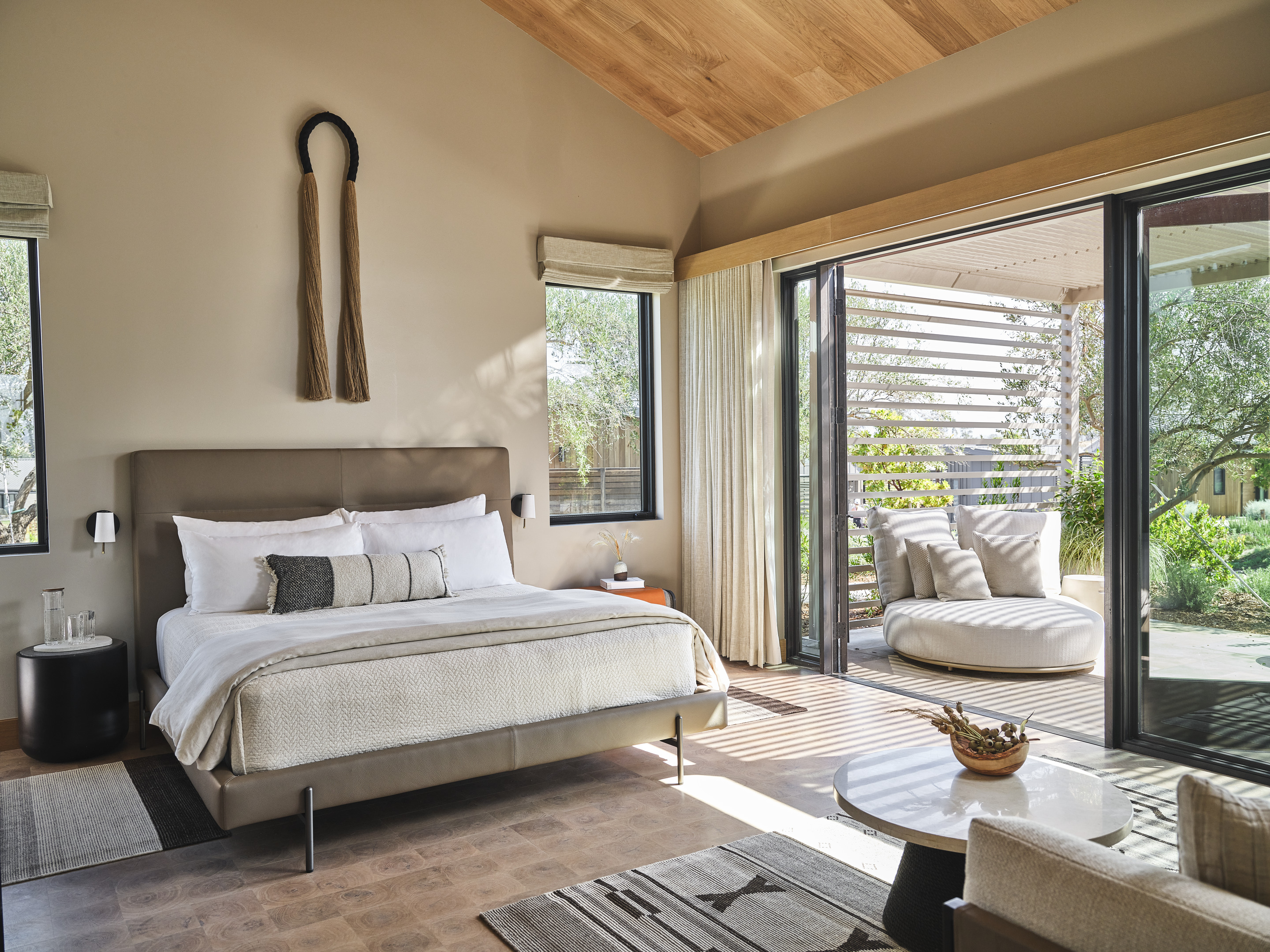 Modern bedroom with a large bed, bedside tables, and decorative wall art from the Auberge Collection. Sliding glass doors open to an outdoor seating area with a cushioned lounge chair. Neutral-toned decor and natural light reminiscent of Stanly Ranch elegance.