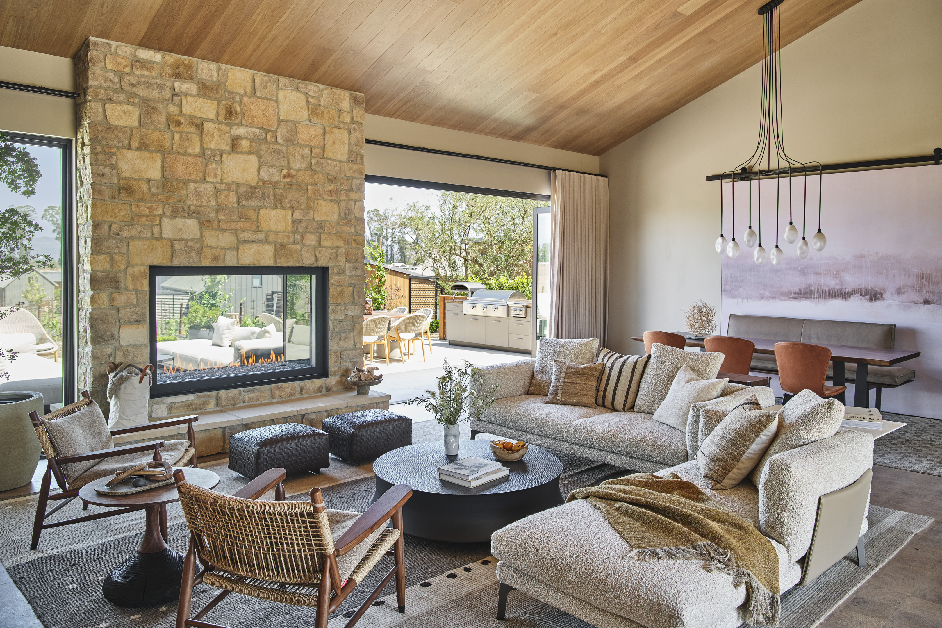 A modern living room with a stone fireplace, comfortable seating, and a view of an outdoor patio mirrors the elegance found in Auberge Resorts. The room features a high ceiling, large windows, and sleek decor elements reminiscent of the luxurious style of Stanly Ranch.