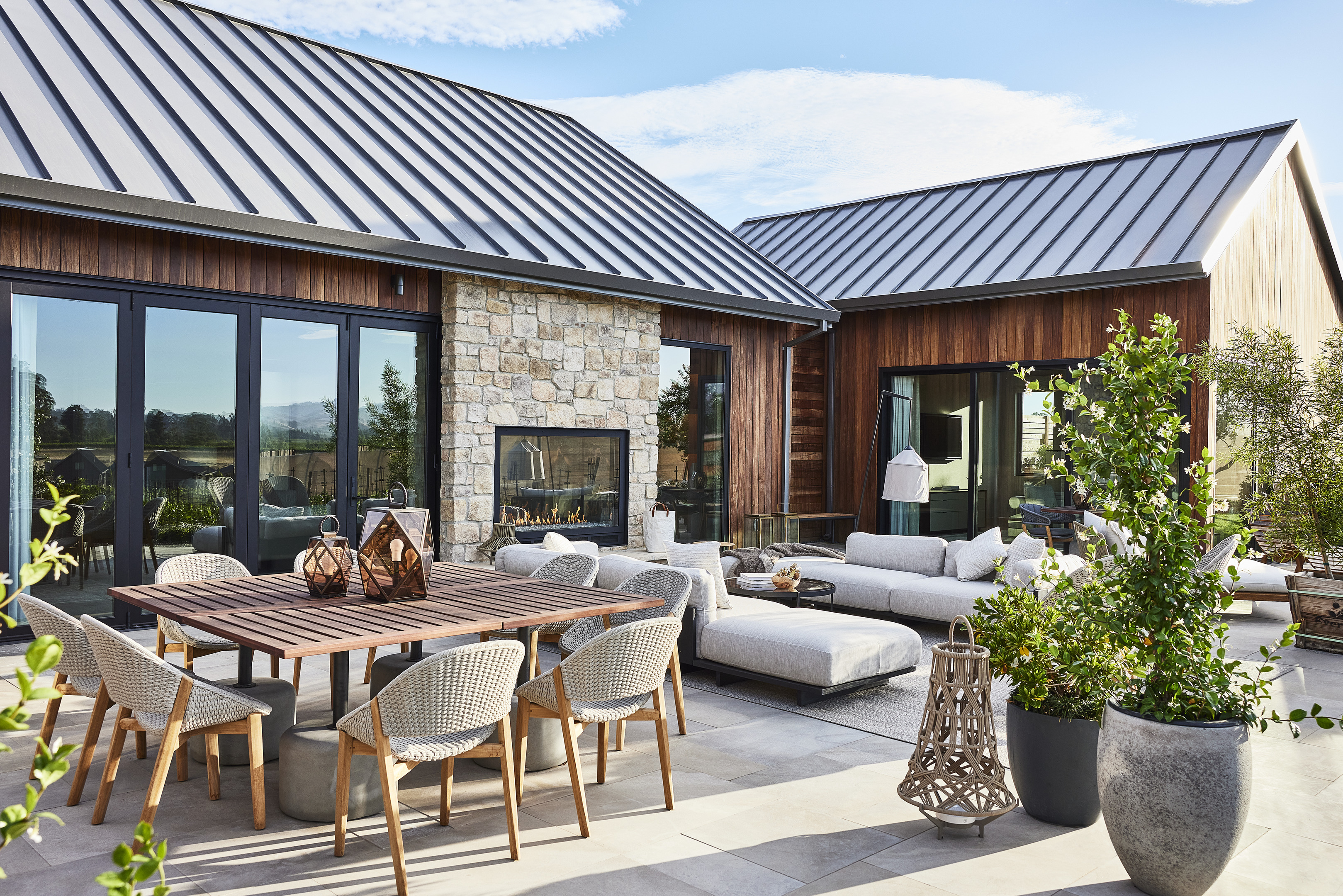 Outdoor patio with wooden dining table and chairs, white sectional sofa, potted plants, and a stone fireplace. Modern buildings with metal roofs surround the patio. The sky is clear, and the weather is sunny—a perfect setting reminiscent of luxury resorts like Auberge Resorts' Stanly Ranch.