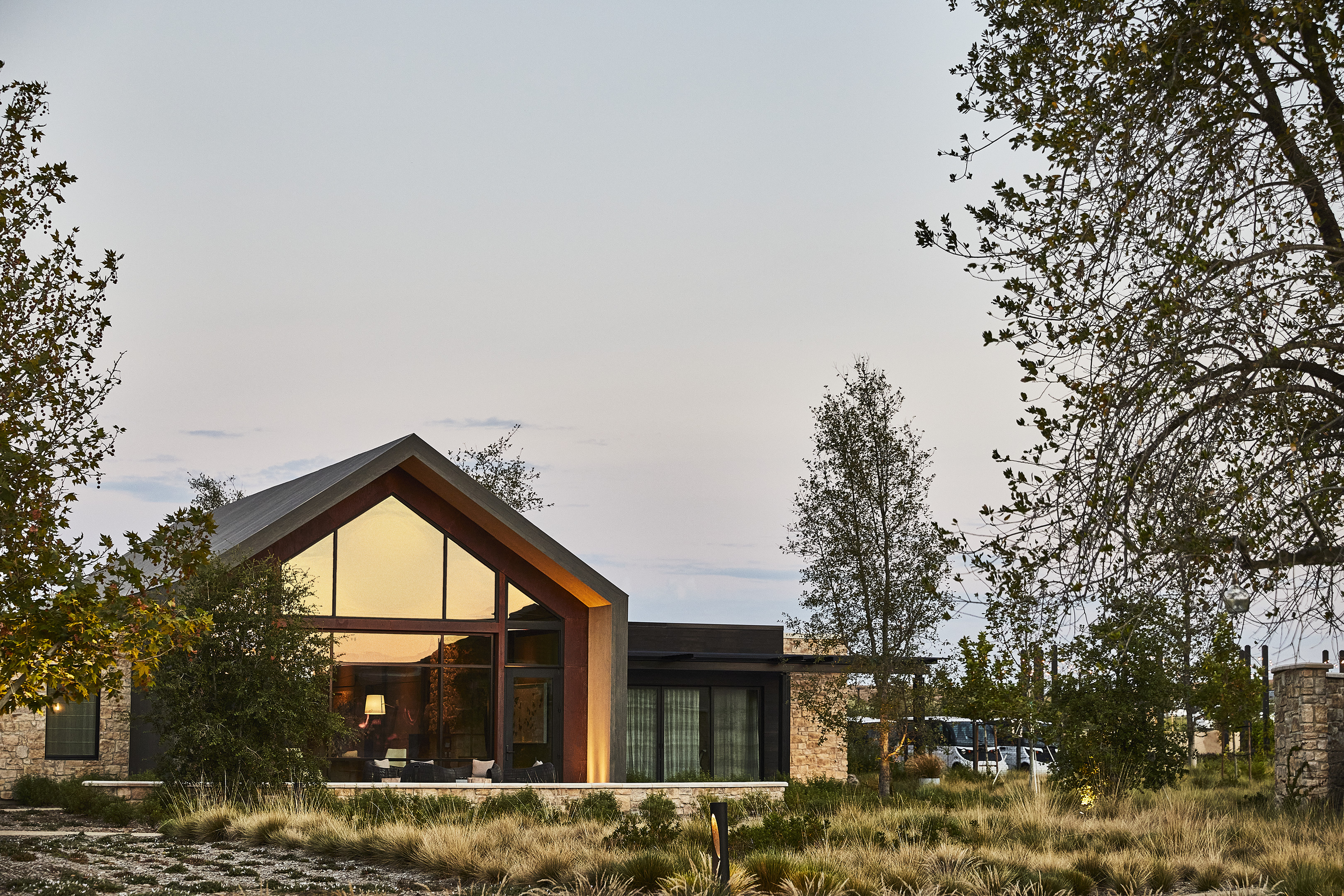 A modern house with large glass windows and wooden accents is surrounded by trees and dry grassland under a cloudy sky, reminiscent of the serene elegance found at Stanly Ranch, part of the Auberge Resorts Collection.