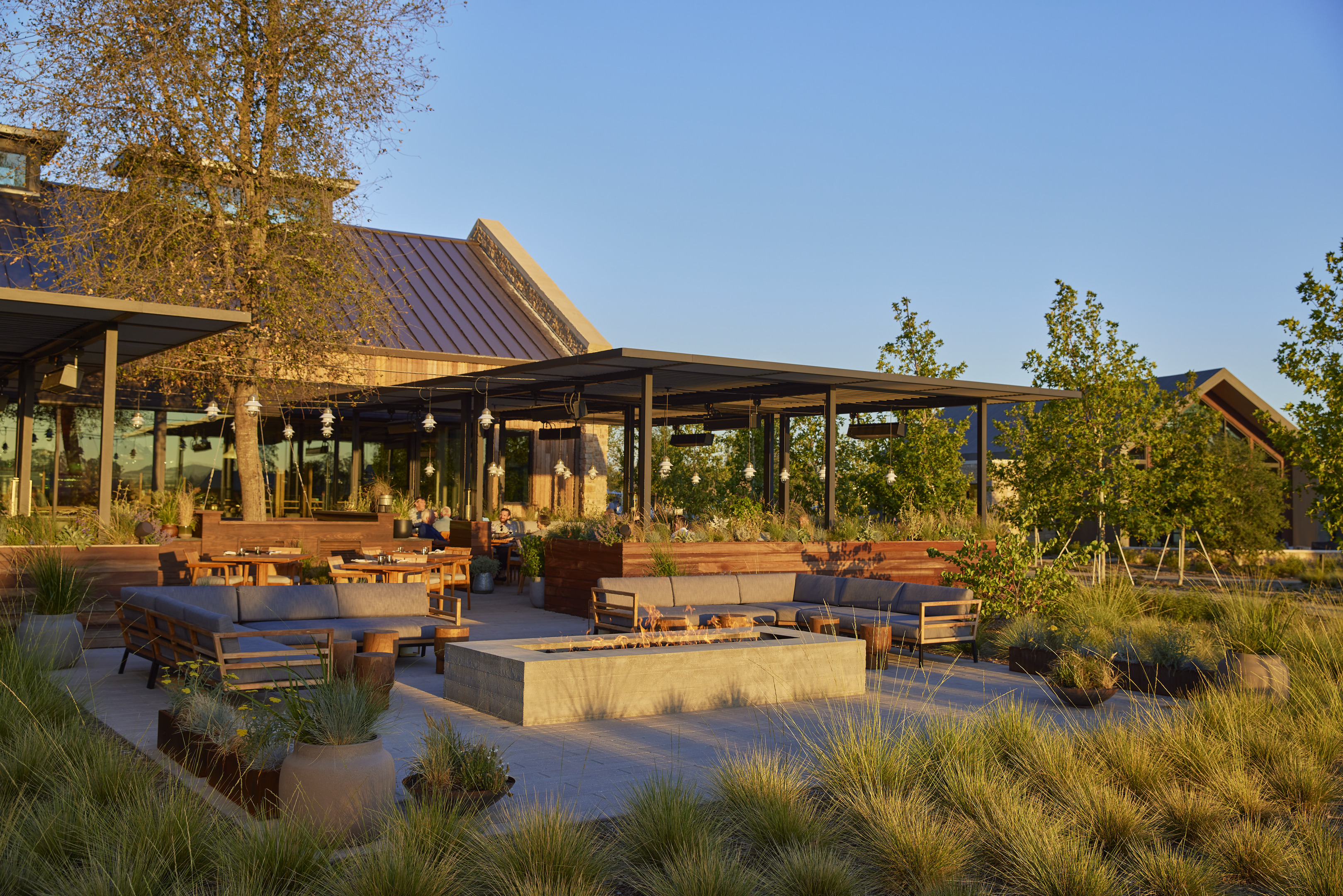 An outdoor seating area with a mix of modern and rustic furniture surrounded by greenery, featuring a fire pit in the center and a covered patio in the background under a clear sky, reminiscent of the luxurious ambiance found at Stanly Ranch, part of the Auberge Resorts Collection.