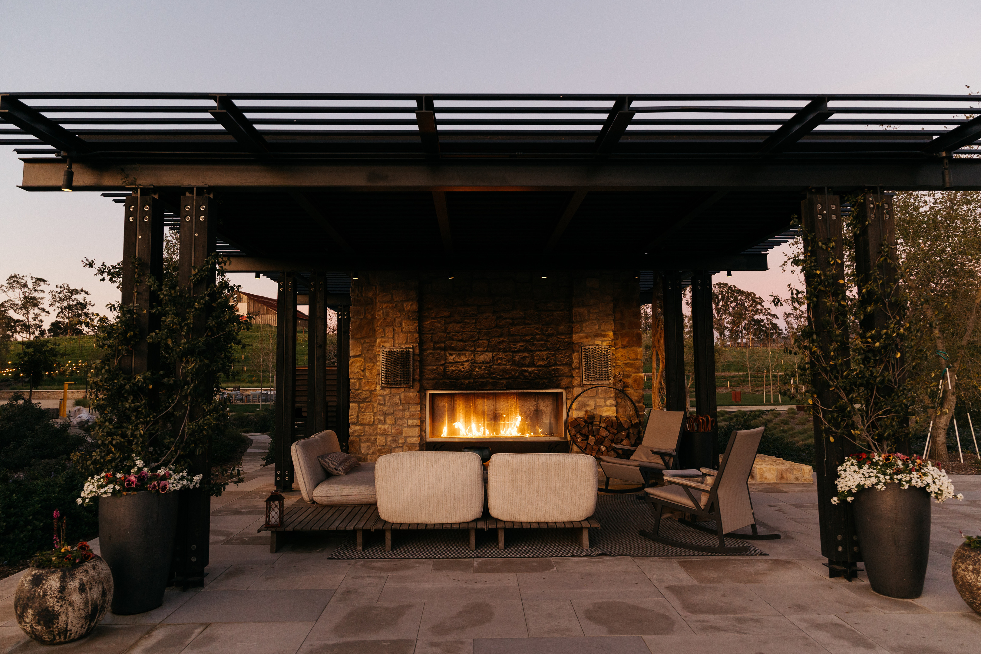 Outdoor patio with a pergola, stone fireplace, and cushioned seating at Stanly Ranch, an Auberge Resort. Planters with flowers surround the area, and a rural landscape is visible in the background at dusk.