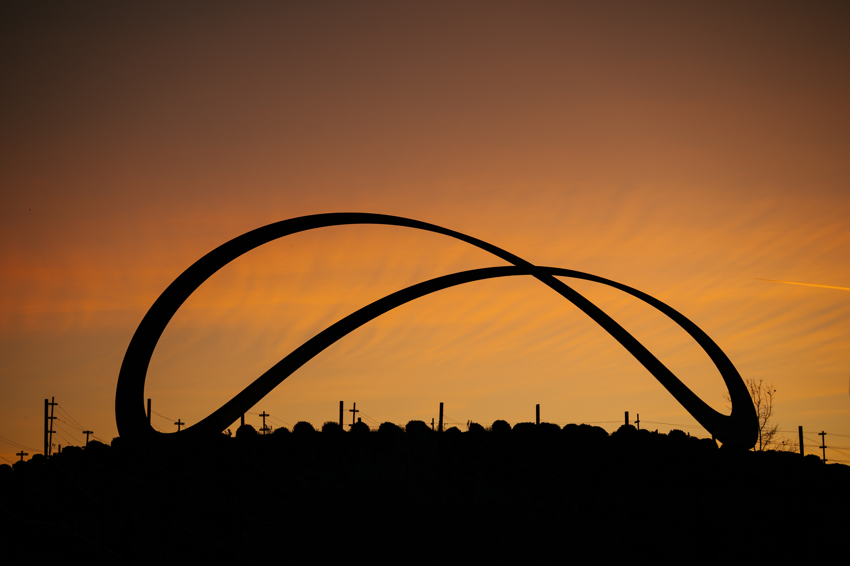 A large, curved metal sculpture silhouetted against an orange and yellow sunset sky, with a few visible utility poles and wires in the background. This serene setting is akin to the picturesque views at Stanly Ranch, part of the Auberge Resorts Collection.