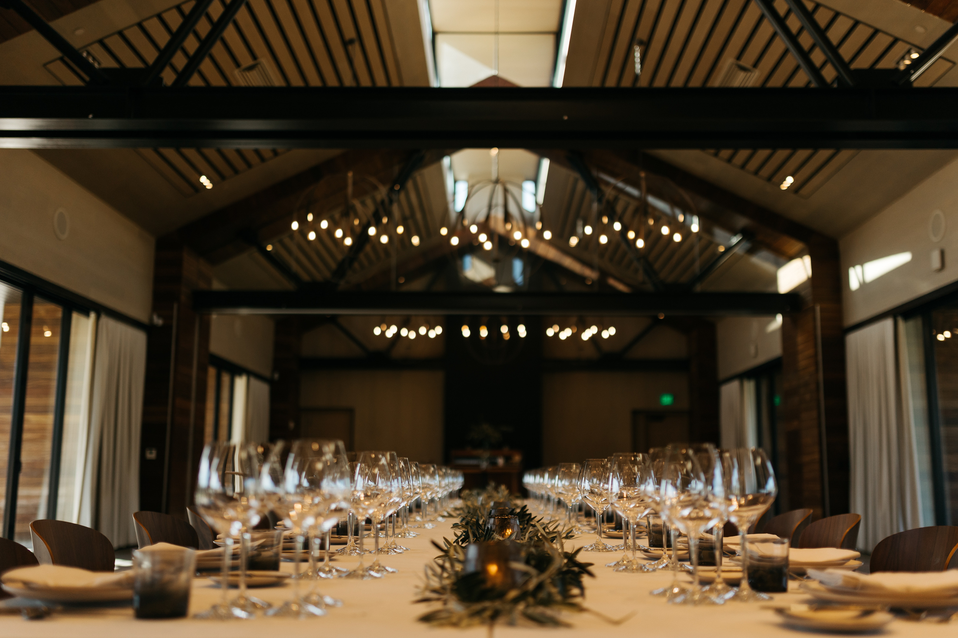 A long dining table set with wine glasses, plates, and cutlery under warm lighting in a room with wooden beams and large windows at Stanly Ranch, Auberge Resorts Collection.