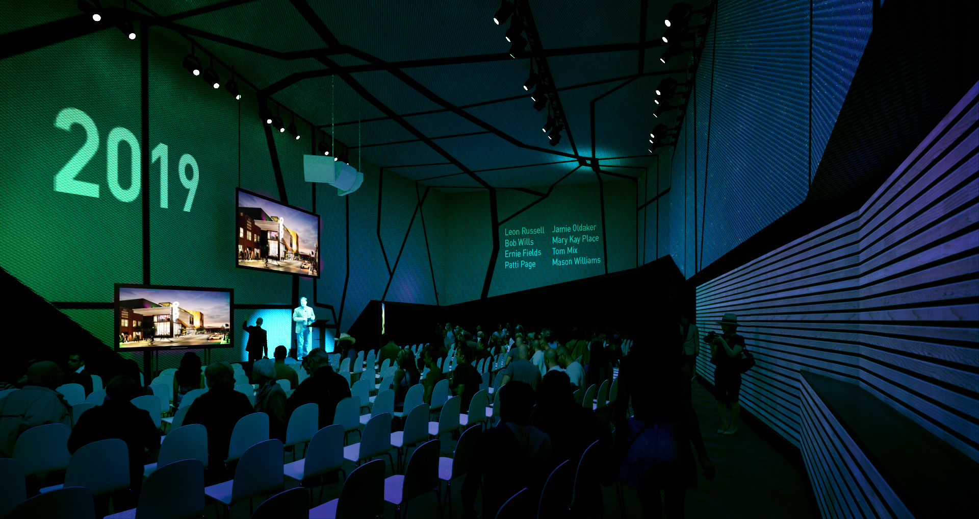 A conference room with a large screen displaying "2019" and presentation slides. Attendees sit facing the stage, where a speaker is presenting about the Oklahoma Museum of Popular Culture (OkPOP). The room has a modern, dark-lit design with blue and green lighting.