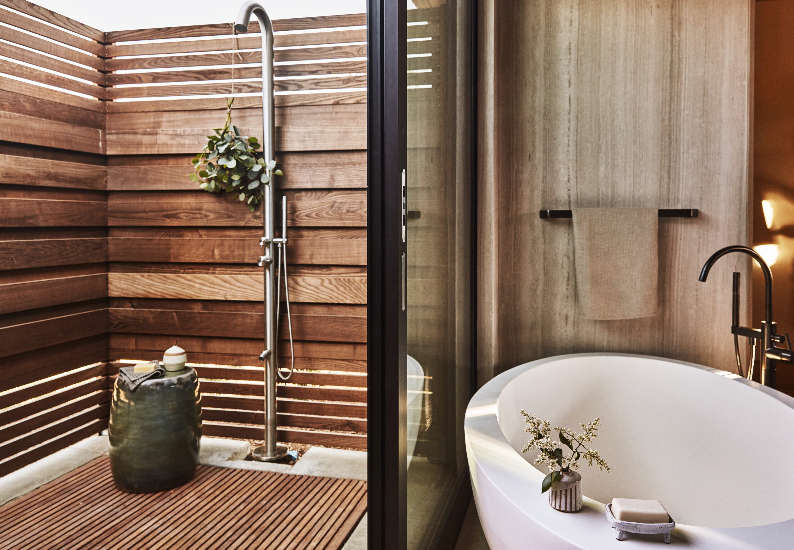 Modern bathroom with a freestanding tub and outdoor shower area. The bathroom, inspired by Stanly Ranch aesthetics, features wooden accents, a black faucet, a rolled towel on a rack, and potted plants for decor.