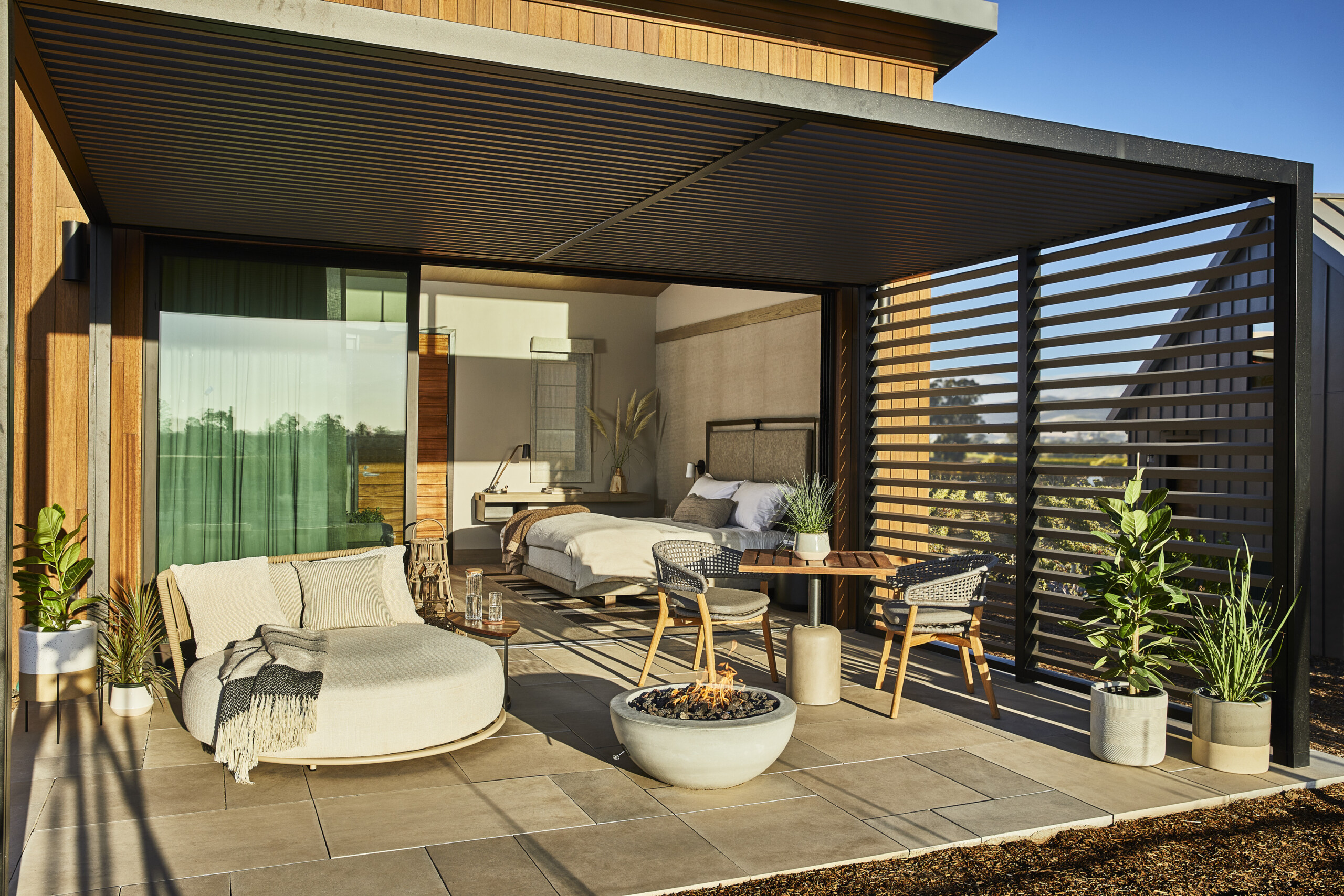 Outdoor patio with modern furniture, including a round daybed, fire pit, dining set, and potted plants, adjacent to a bedroom with a large glass door in the Stanly Ranch style.