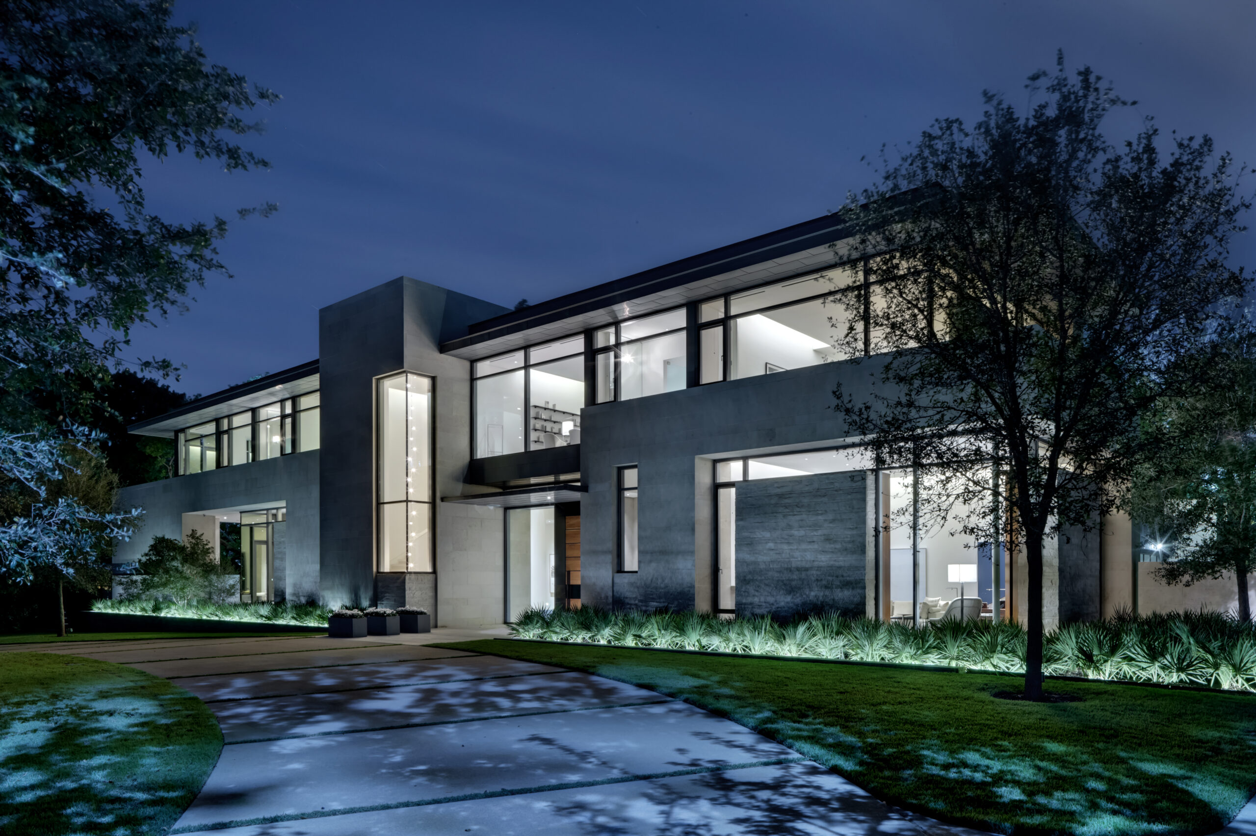 A modern two-story house with extensive glass windows and a well-lit exterior, surrounded by trees and greenery, viewed at night.