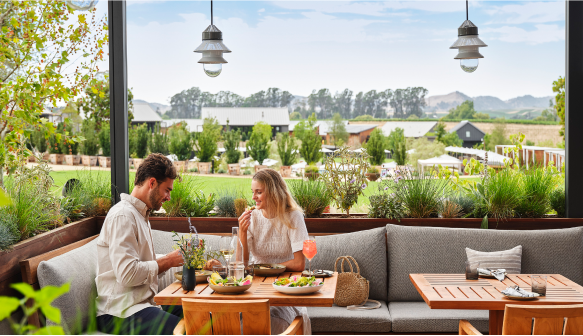 A man and a woman are dining outdoors at Stanly Ranch, enjoying salads and drinks at a wooden table. The scenic view of fields and buildings in the background adds to the charm as they are surrounded by greenery.