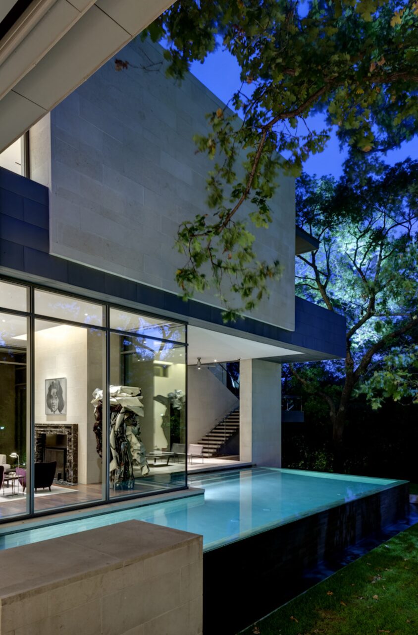 Modern house with large glass windows, a visible staircase inside, and an artwork displayed; exterior features an adjacent reflective pool, trees around, and evening sky in the background.