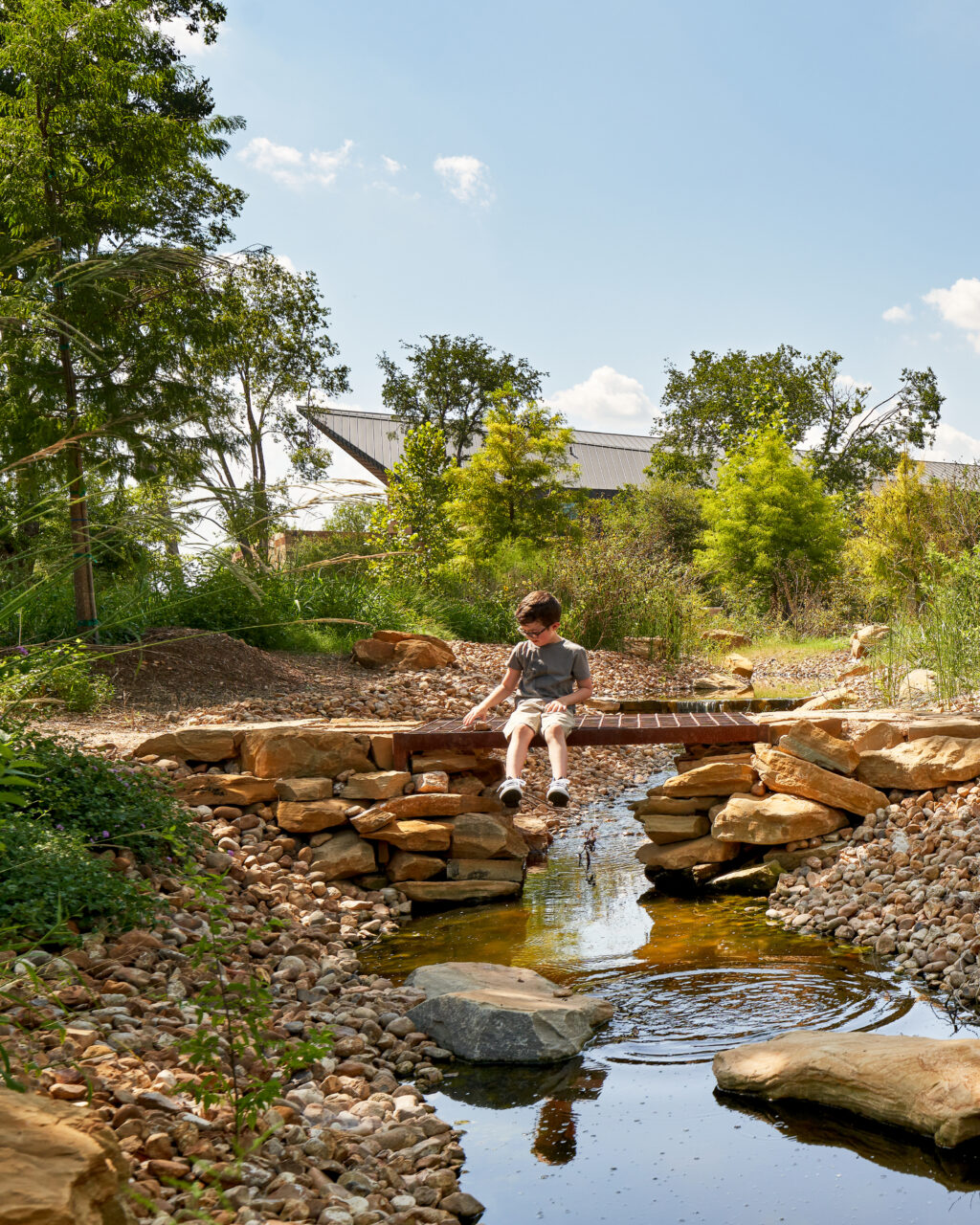 A child sits on a small wooden bridge over a pond in a garden with rocks and green trees under a clear blue sky.