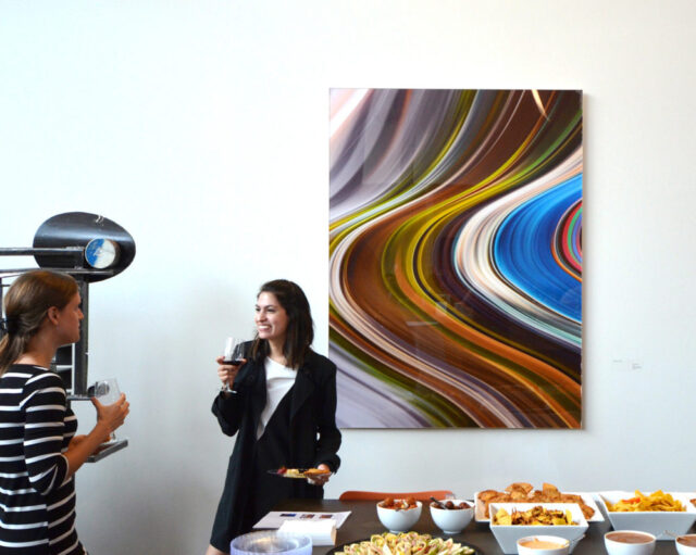 Two people stand with drinks near a table of food, in front of a colorful, abstract painting on a white wall.