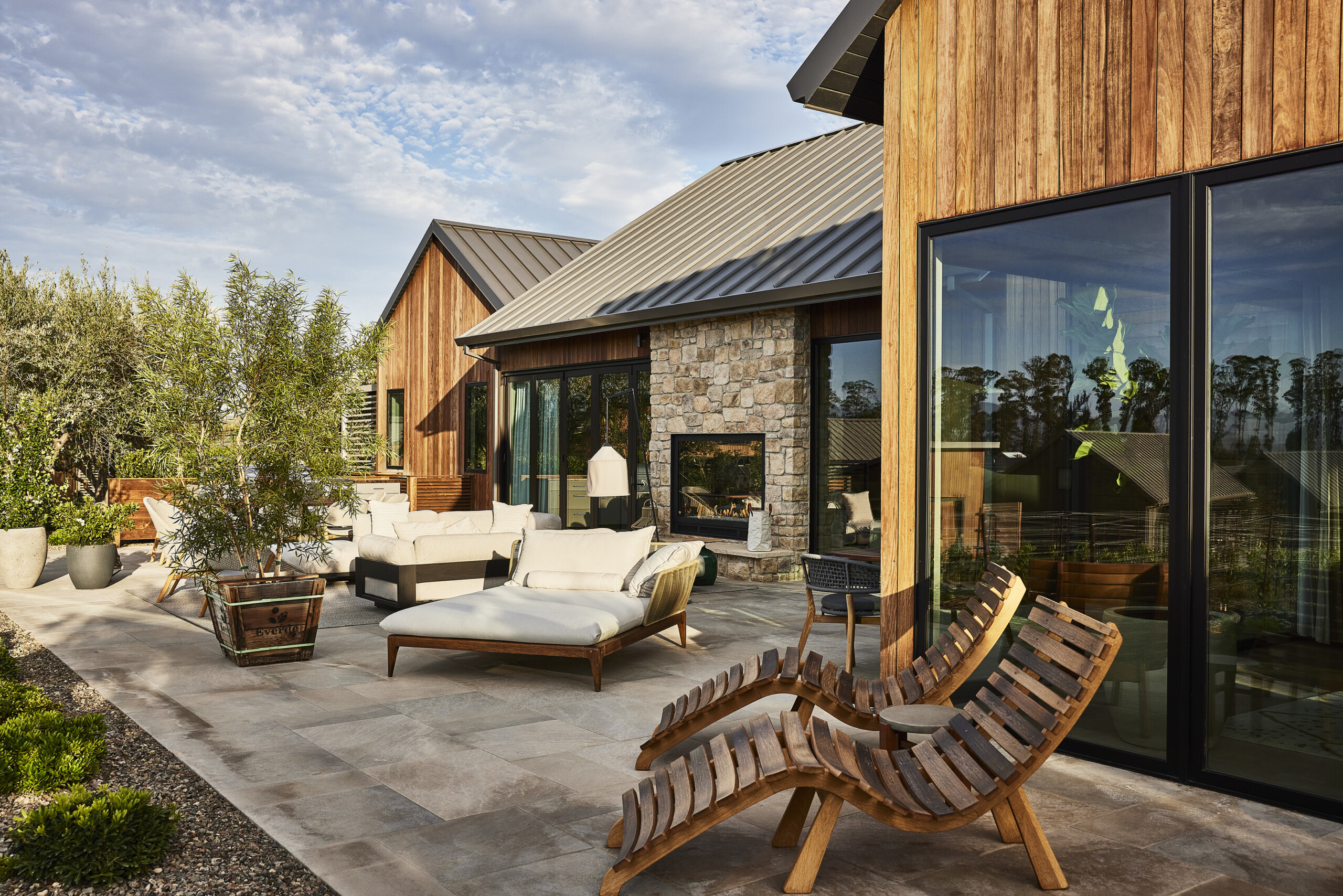 A spacious patio with modern outdoor furniture, including wooden loungers, a couch, and a stone fireplace. The patio is part of a house at Stanly Ranch with wooden and stone exterior walls.