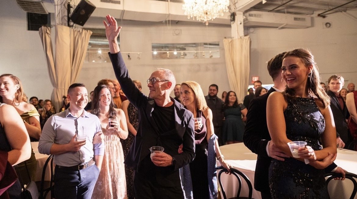 A group of people stand together in a well-lit room. One man is raising his hand while others smile and hold drinks. A chandelier hangs from the ceiling.