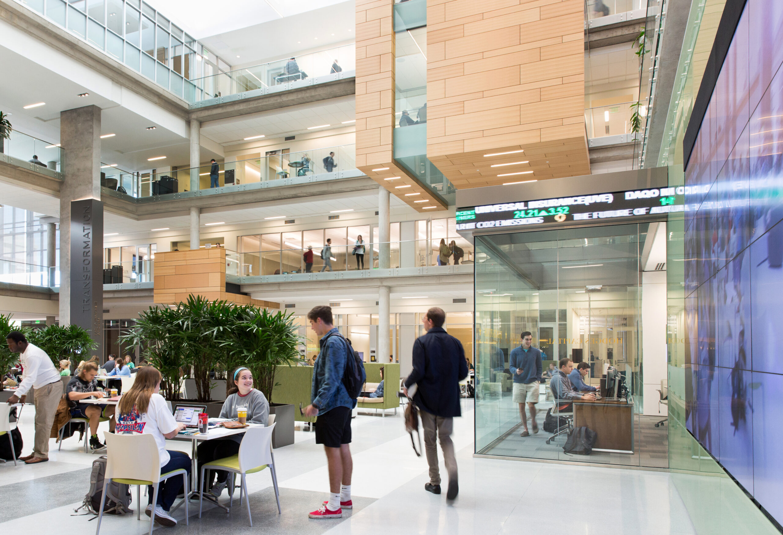 Students and professionals interact and work in a multi-level modern atrium with glass walls, indoor greenery, and large digital screens displaying stock market data.