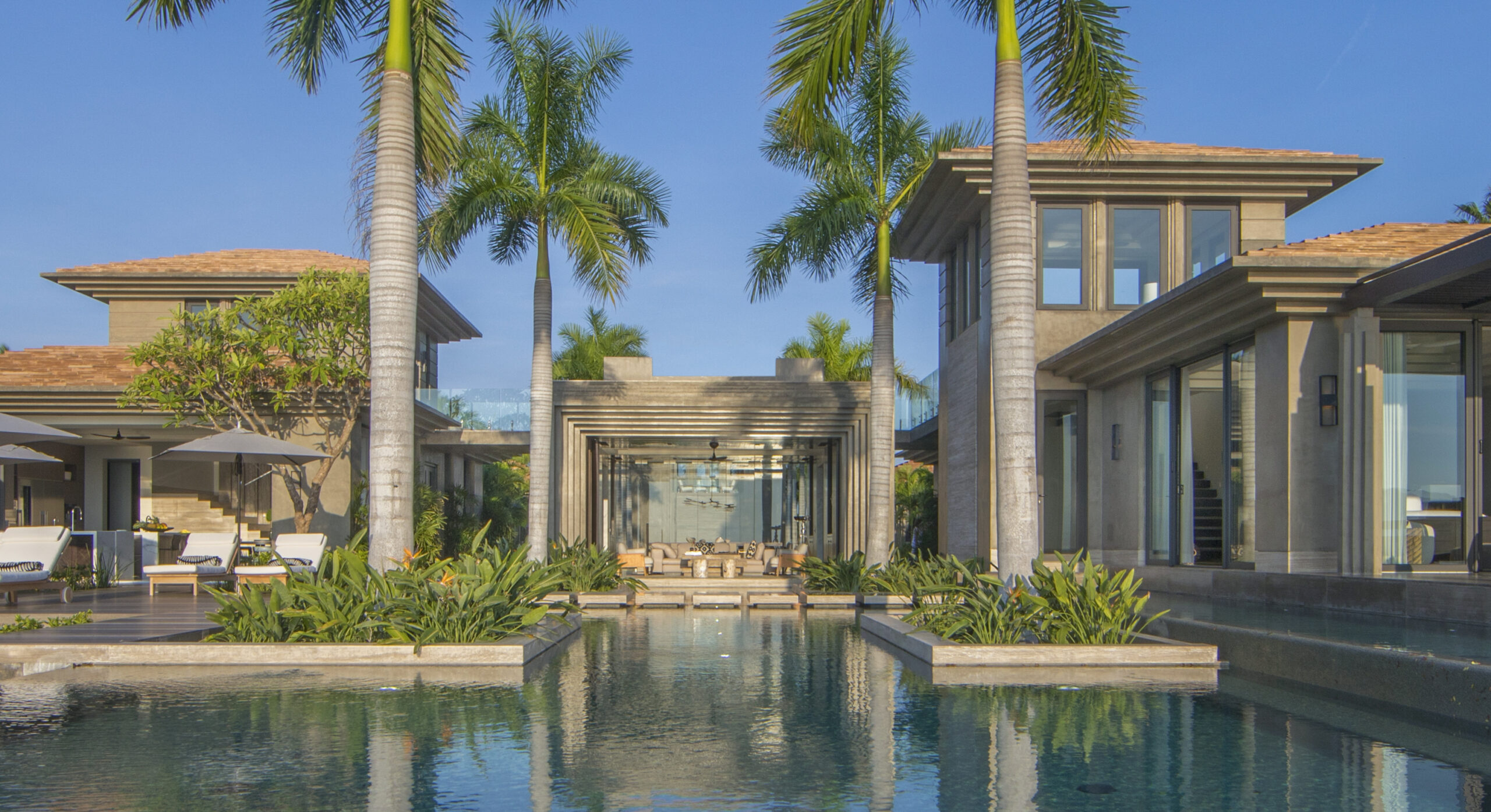 Modern luxury home with a central pool, surrounded by palm trees and lush greenery. Two-story structures with large glass windows and wooden roof shingles are visible against a blue sky backdrop.