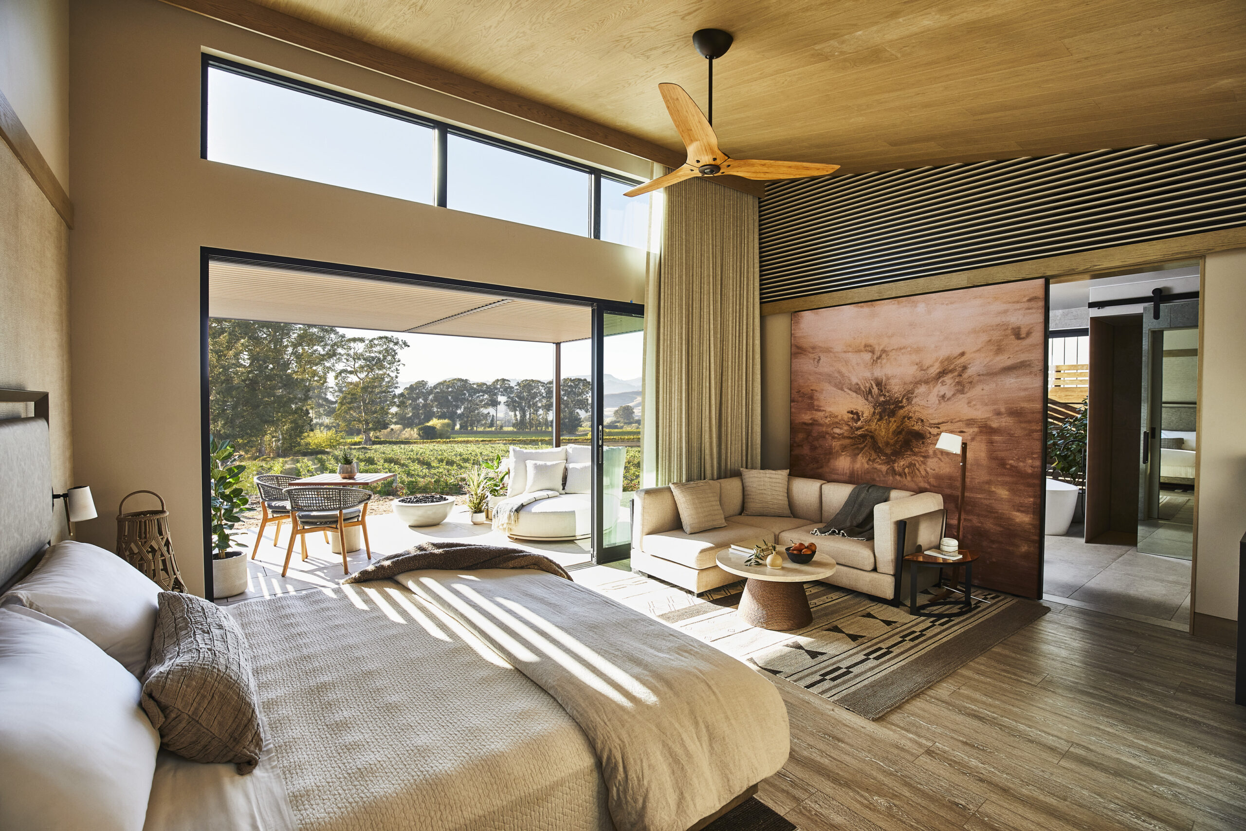 Modern bedroom with a large bed, sitting area, and sliding glass doors leading to an outdoor patio with seating and a view of trees. The room features wooden accents and natural light.