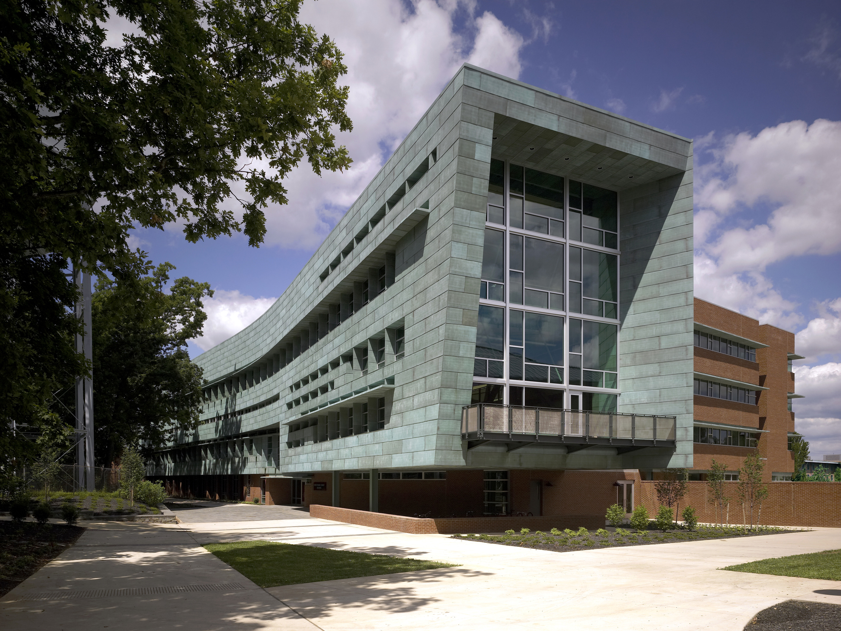 Modern multi-story building with a curved metal facade and large windows, surrounded by trees and a paved walkway, under a partly cloudy sky. This stunning example of architecture can be found at the Stuckeman School at Penn State.