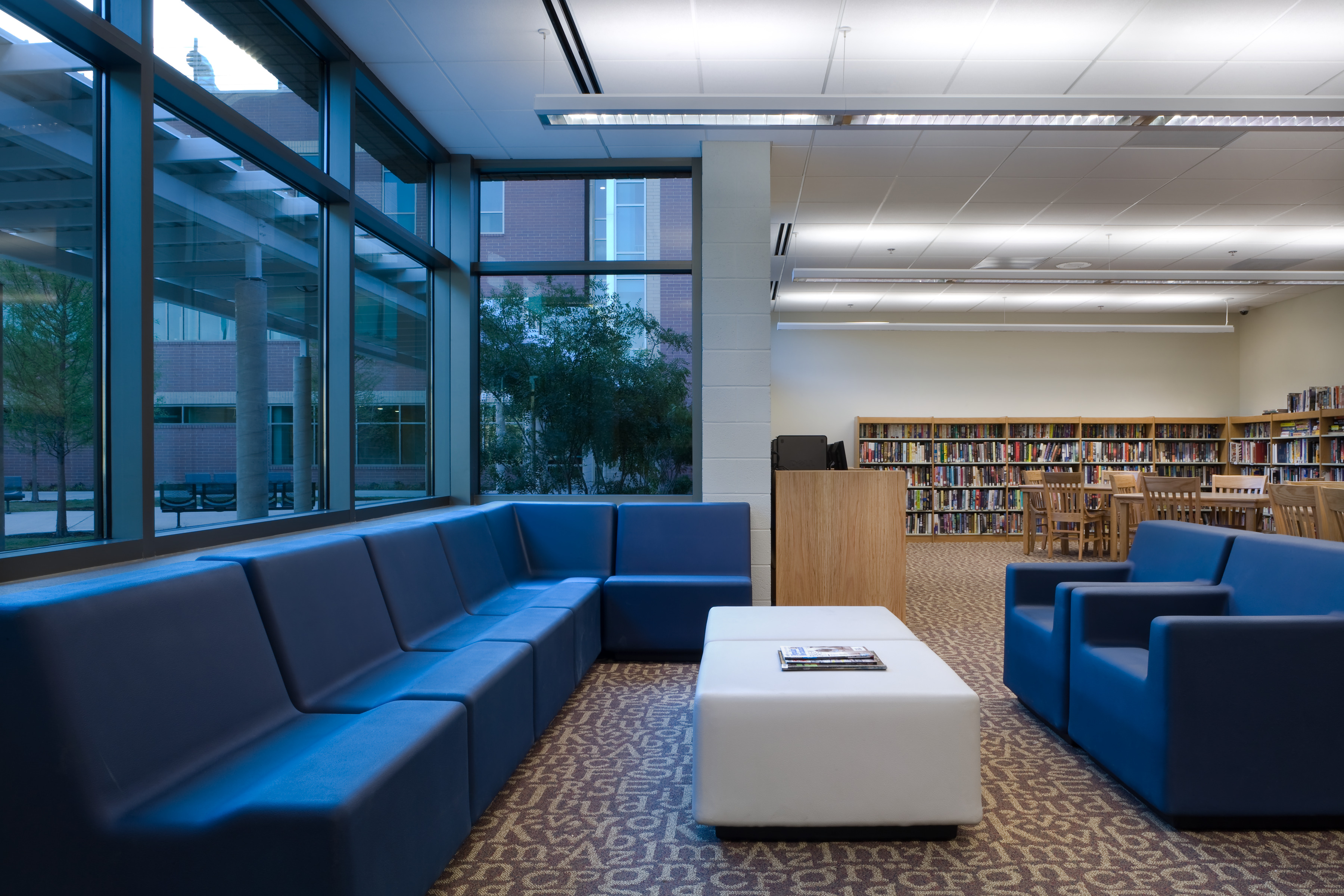 A modern library interior with blue seating, a white coffee table, ceiling lights, large windows, bookshelves, and a few wooden tables and chairs creates a welcoming atmosphere reminiscent of a recovery center.