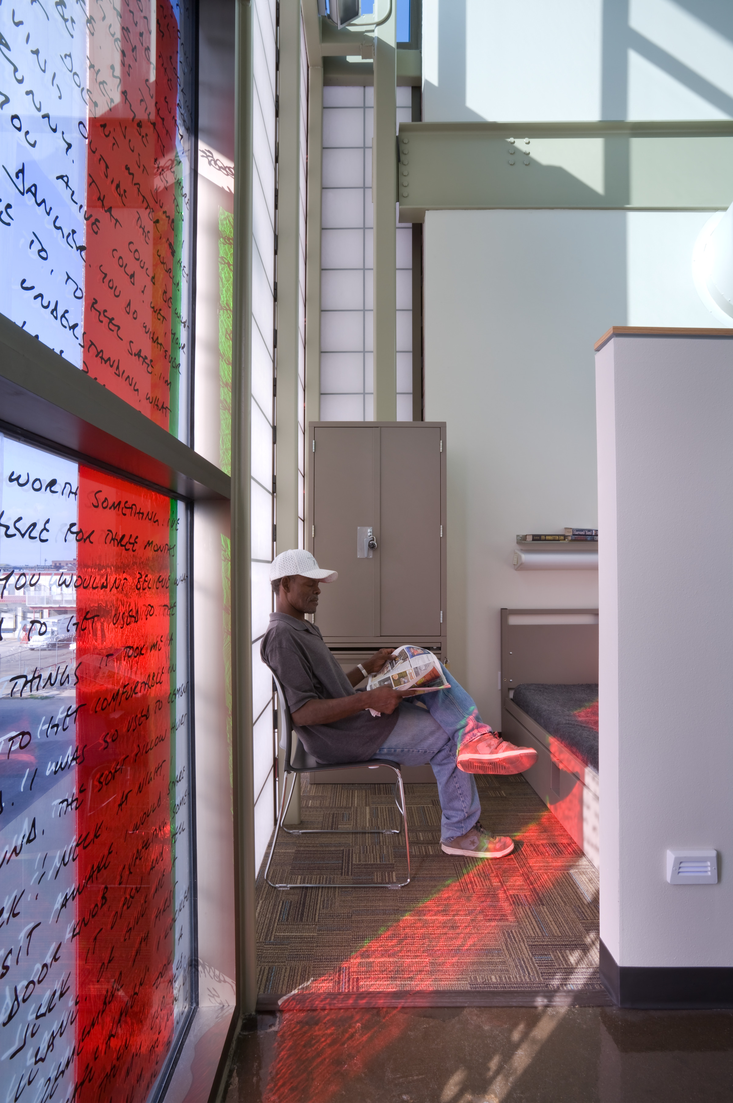 A person sits on a chair near a sunlit window, reading a magazine in a modern recovery center with red and black lines on the glass and various shelves and seating areas.