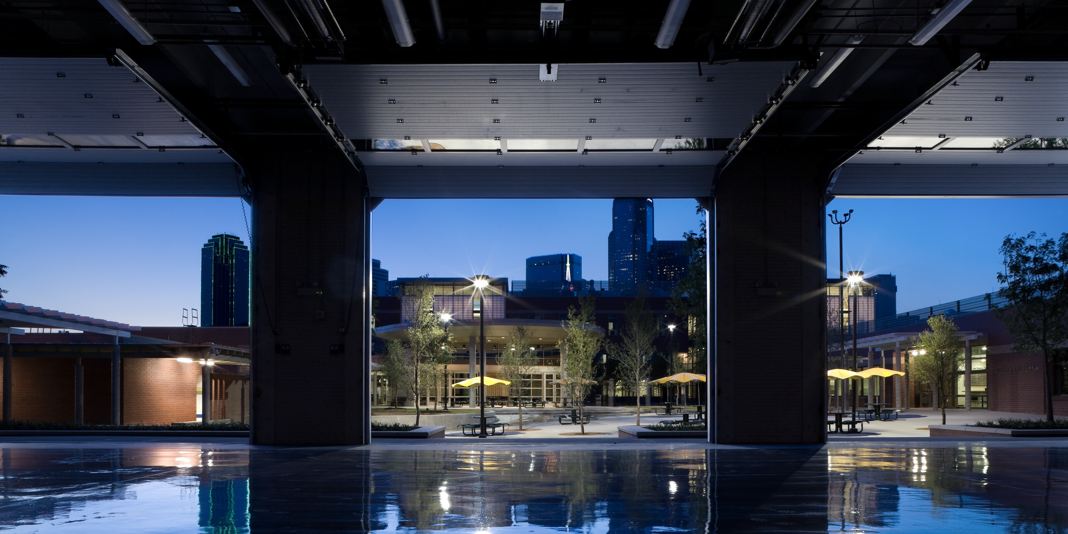 Industrial structure with open garage doors revealing an outdoor area at dusk with benches, yellow umbrellas, and illuminated buildings in the background. Reflective wet floor in the foreground hints at a welcoming atmosphere of a nearby Homeless Recovery Center.