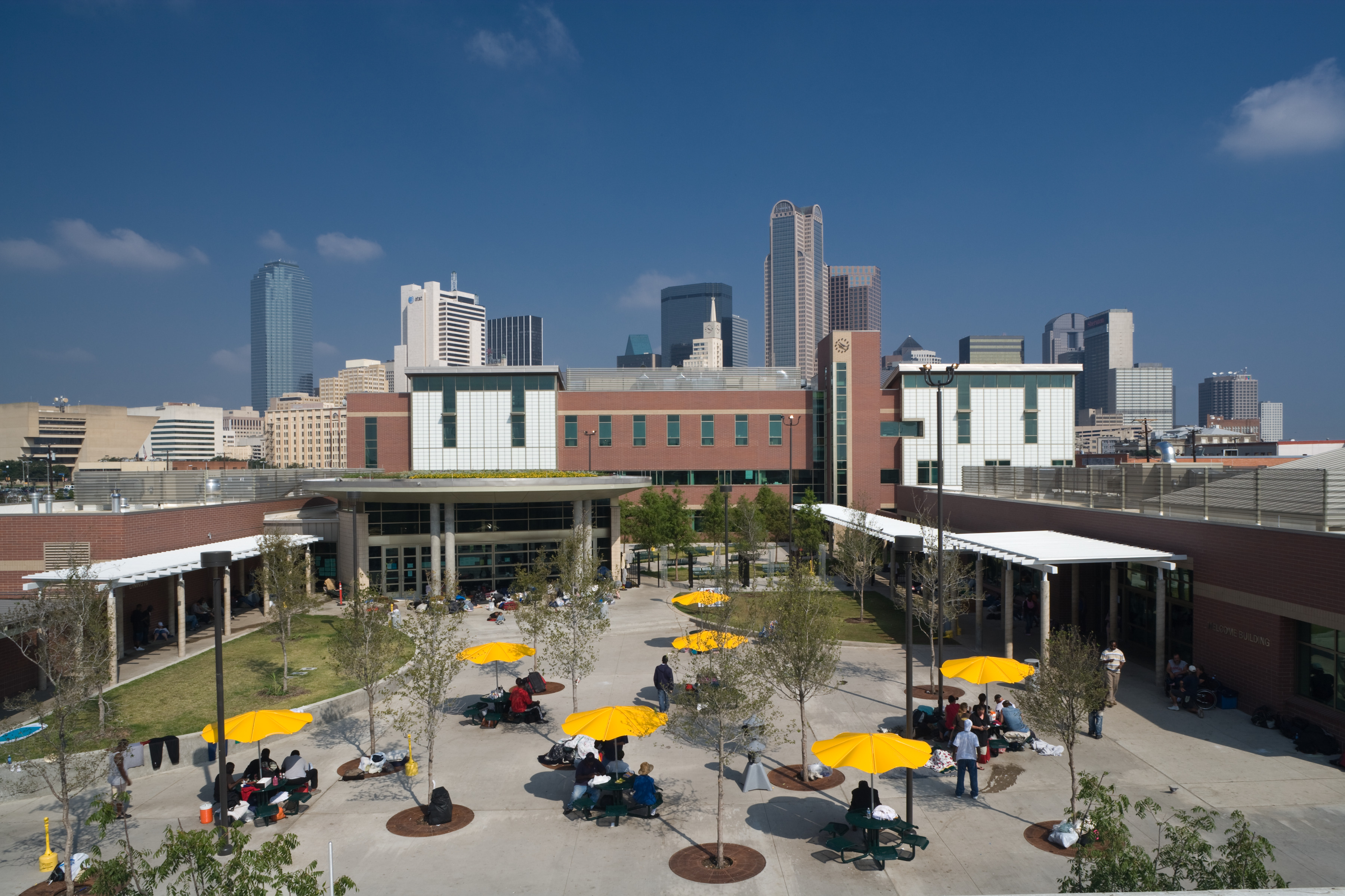 An outdoor courtyard with yellow umbrella tables, people sitting, buildings in the background, and a clear blue sky, near the new bridge to the Homeless Recovery Center.