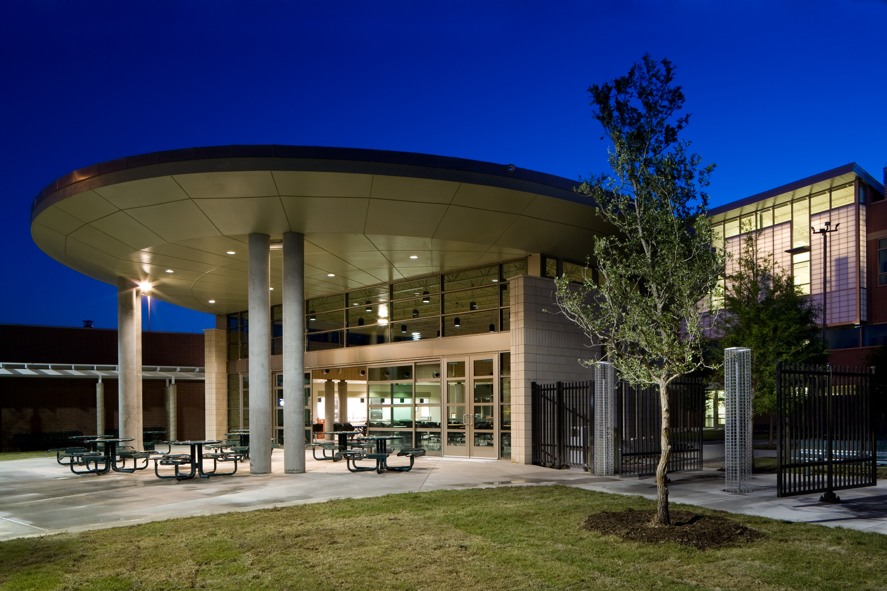 An exterior view of a modern Recovery Center with large columns supporting a circular roof, outdoor seating, and a tree in the foreground, photographed at dusk.