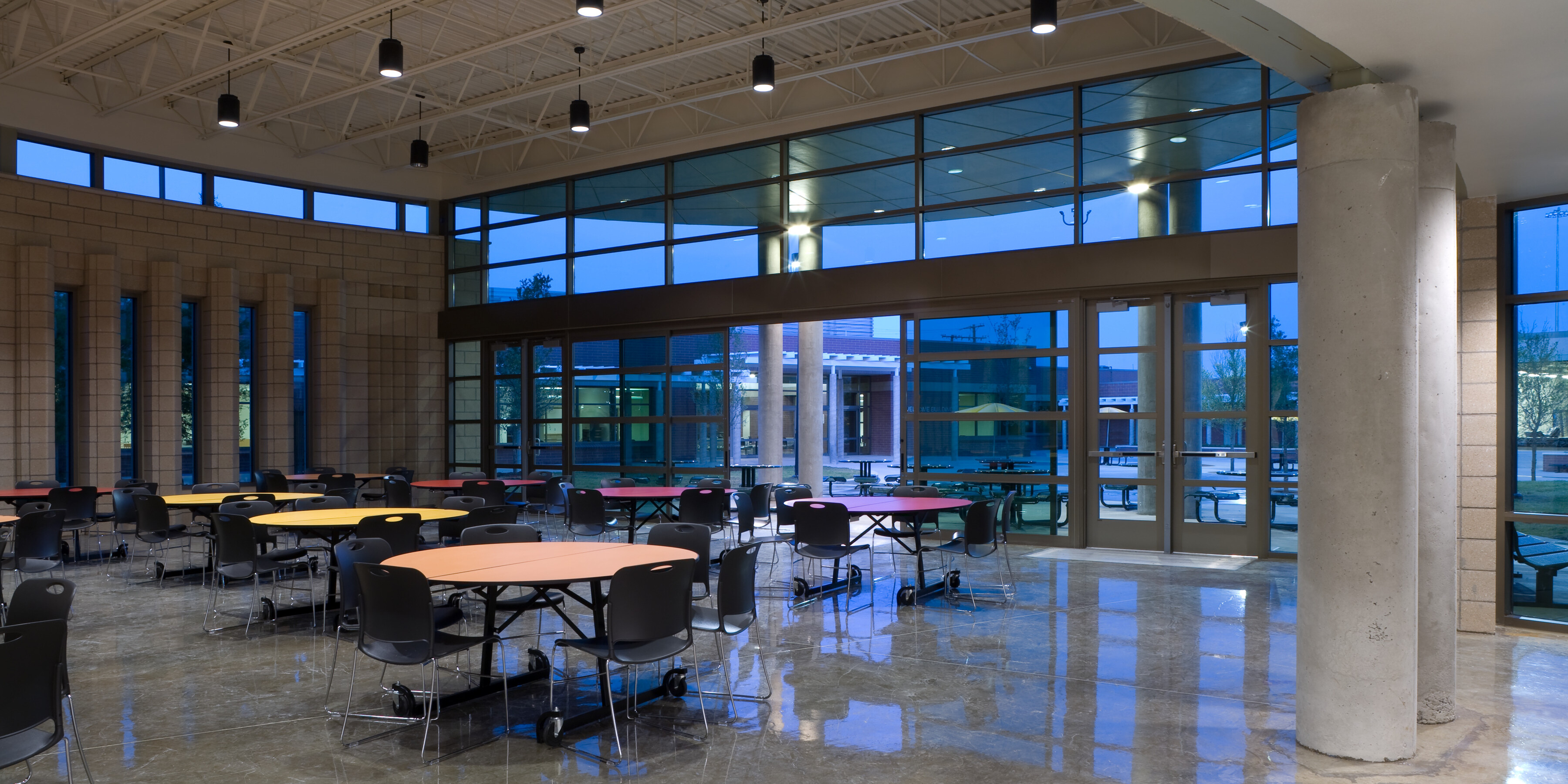 A spacious, modern cafeteria with large windows, round tables, black chairs, and overhead lighting. The polished concrete floor complements the serene view of the outside area. This welcoming space is part of our Homeless Recovery Center.