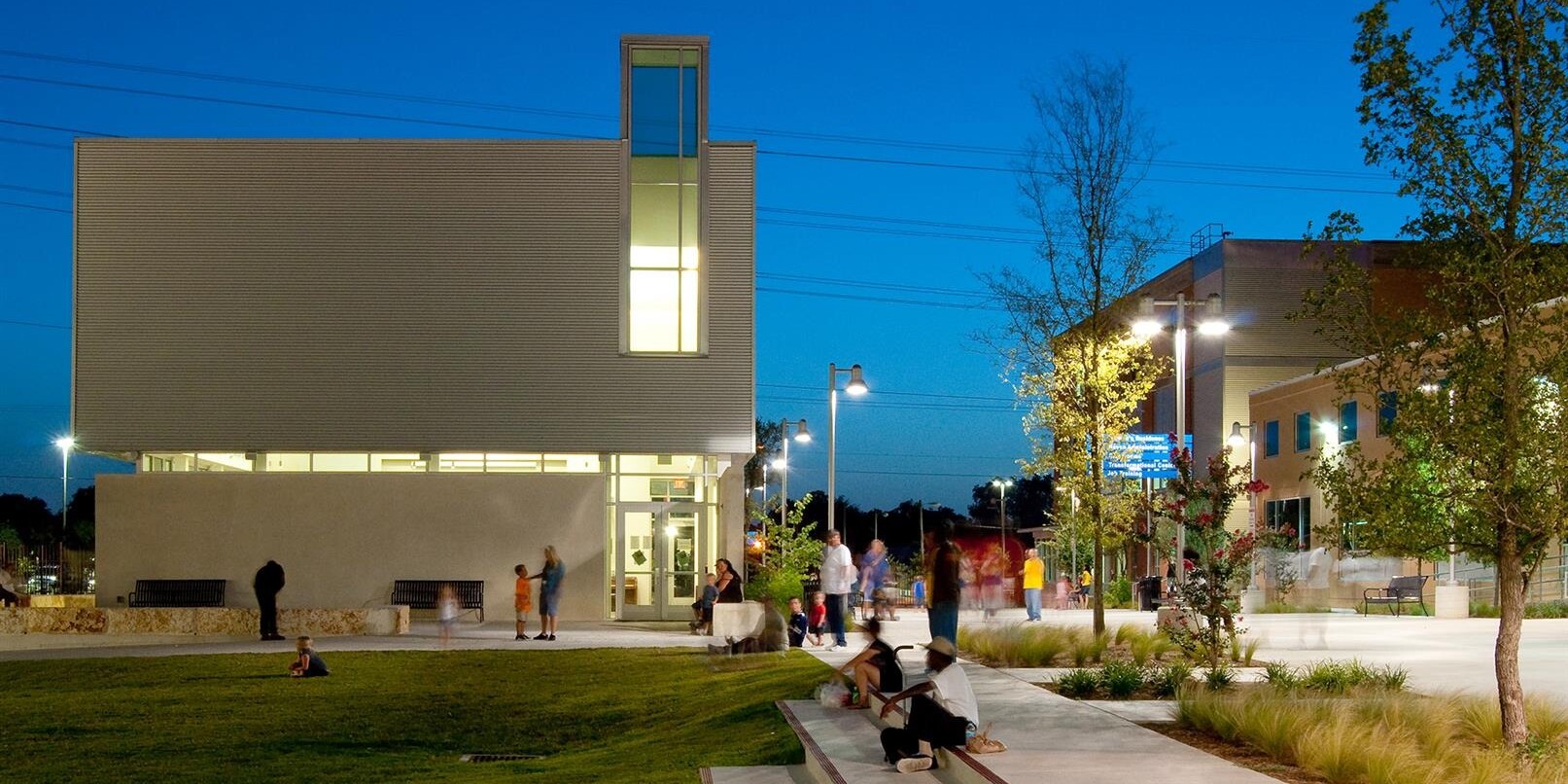 A modern building with large windows is illuminated at dusk, serving as a Haven for Hope. People are walking and sitting in a landscaped area with grass, trees, and concrete pathways.