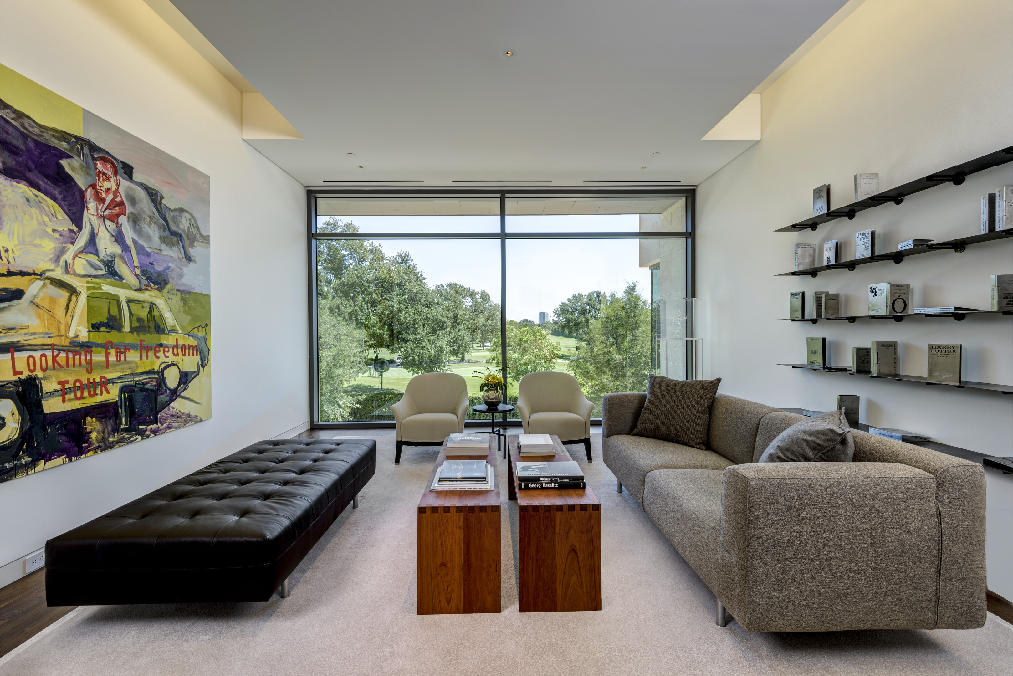 A modern residence featuring a gray sofa, dark leather bench, two wooden coffee tables, floor-to-ceiling windows with a scenic view, wall-mounted shelving filled with books, and vibrant ART showcased through a colorful painting.