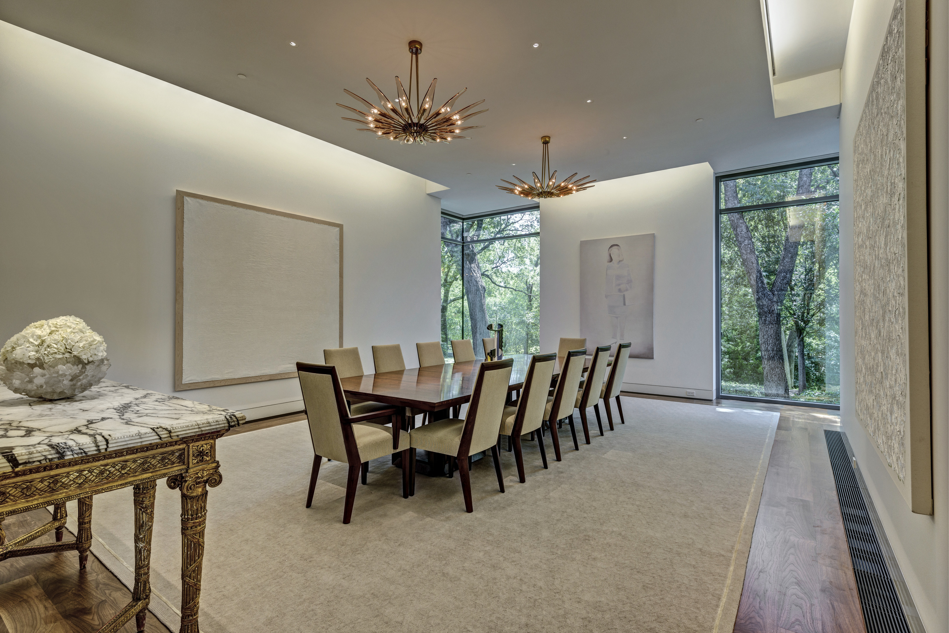 A modern dining room in a luxurious residence featuring a long wooden table with ten upholstered chairs, large abstract artwork, a marble top table, floor-to-ceiling windows, and two starburst chandeliers.