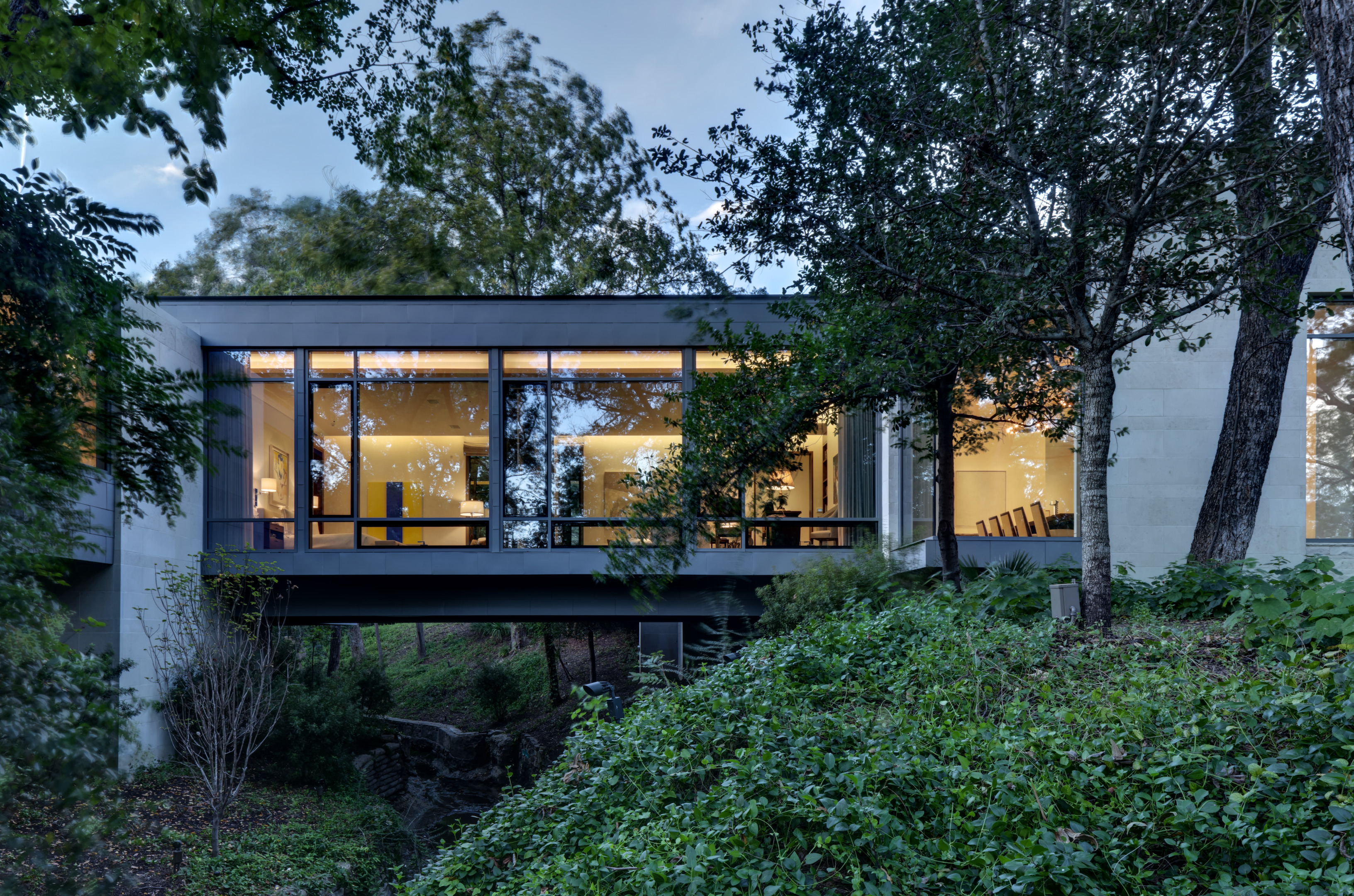 A modern residence with large windows is elevated over a lush, green landscape. Warm lighting inside the building contrasts with the surrounding foliage under a cloudy sky, creating an artful blend of nature and modernity.