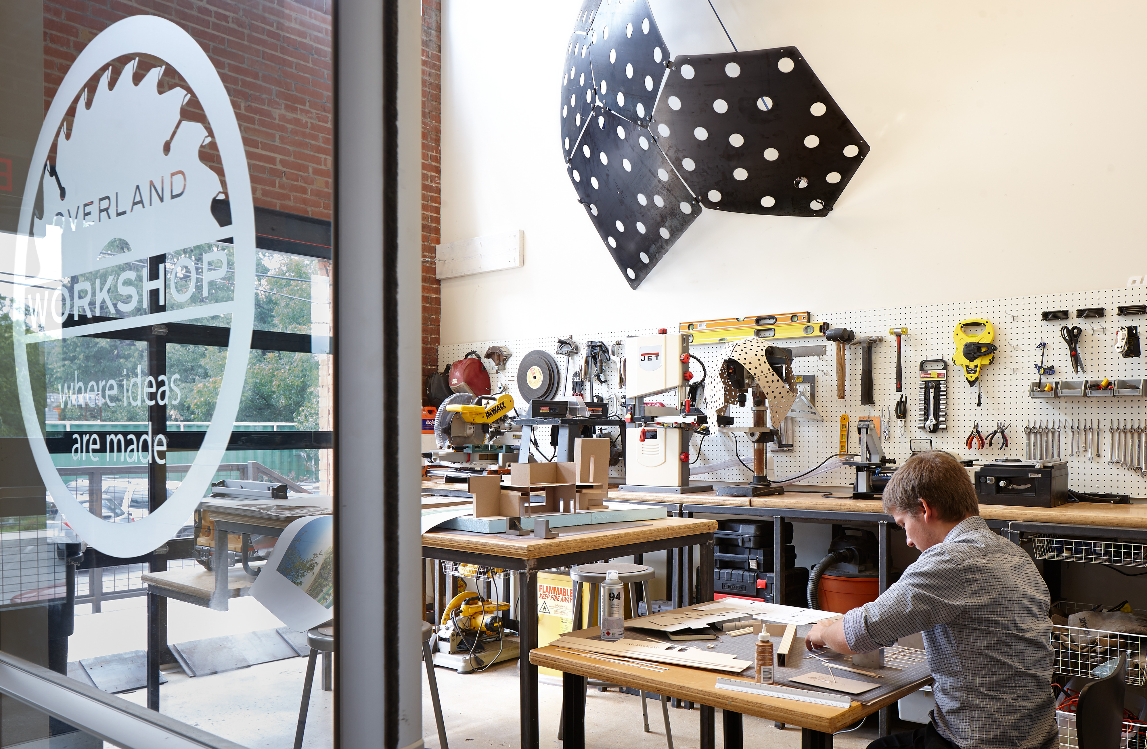 A person works on a project at a cluttered workshop desk, embodying innovation. A large geometrical piece with polka dots hangs on the wall. Various tools and materials are meticulously organized on pegboards and shelves, providing an inspiring backdrop for creative breakthroughs.