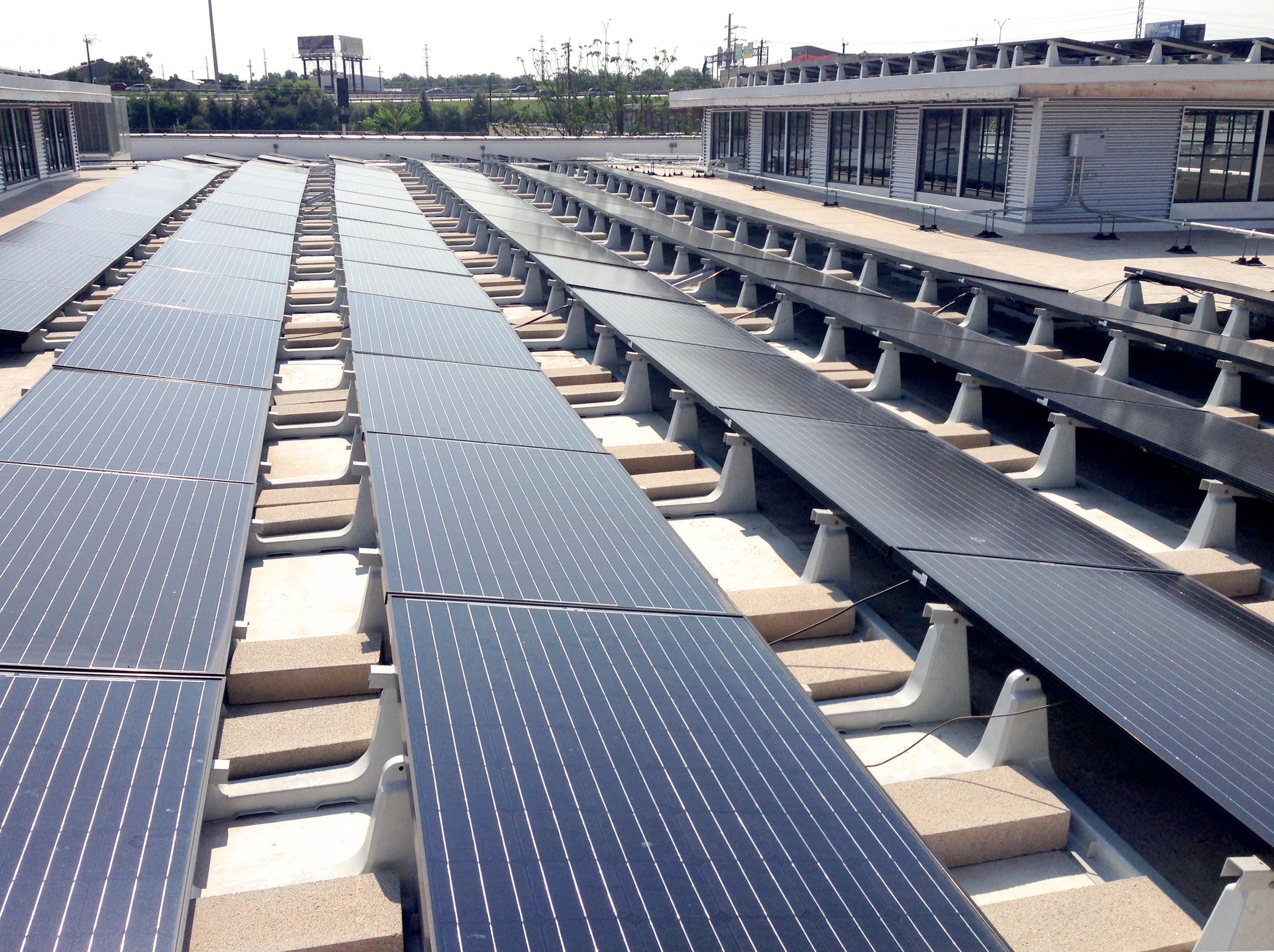 Rows of solar panels installed on a flat rooftop under a clear sky, with industrial buildings like Hughes Warehouse in the background.