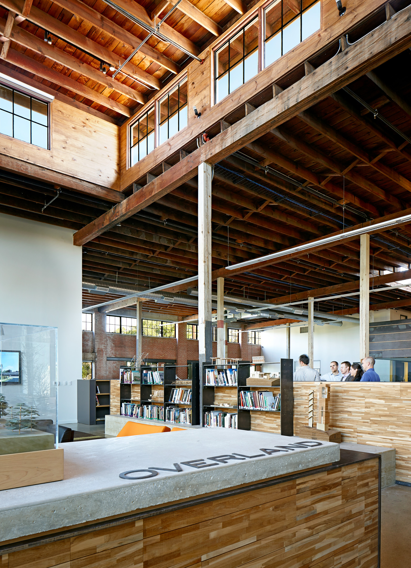 A modern library with wooden beams and high ceilings, shelves filled with books, a reception desk with "OVERLAND" inscribed, reminiscent of the historic Hughes Warehouse, and people conversing in the background.