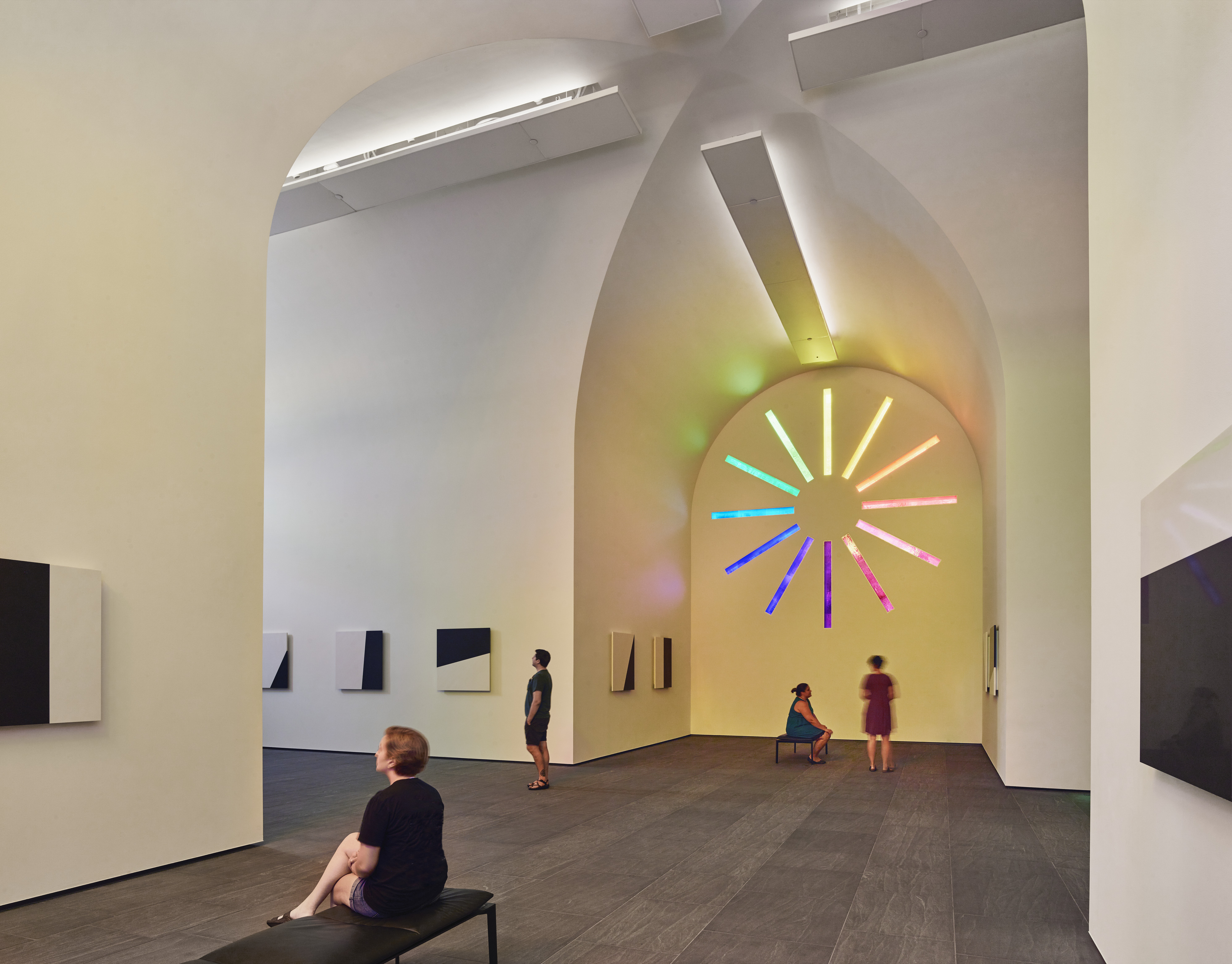 People observe art installations in a modern gallery with high ceilings in Austin. A circular neon artwork with multicolored lights, reminiscent of Ellsworth Kelly's vibrant style, is displayed on the back wall.