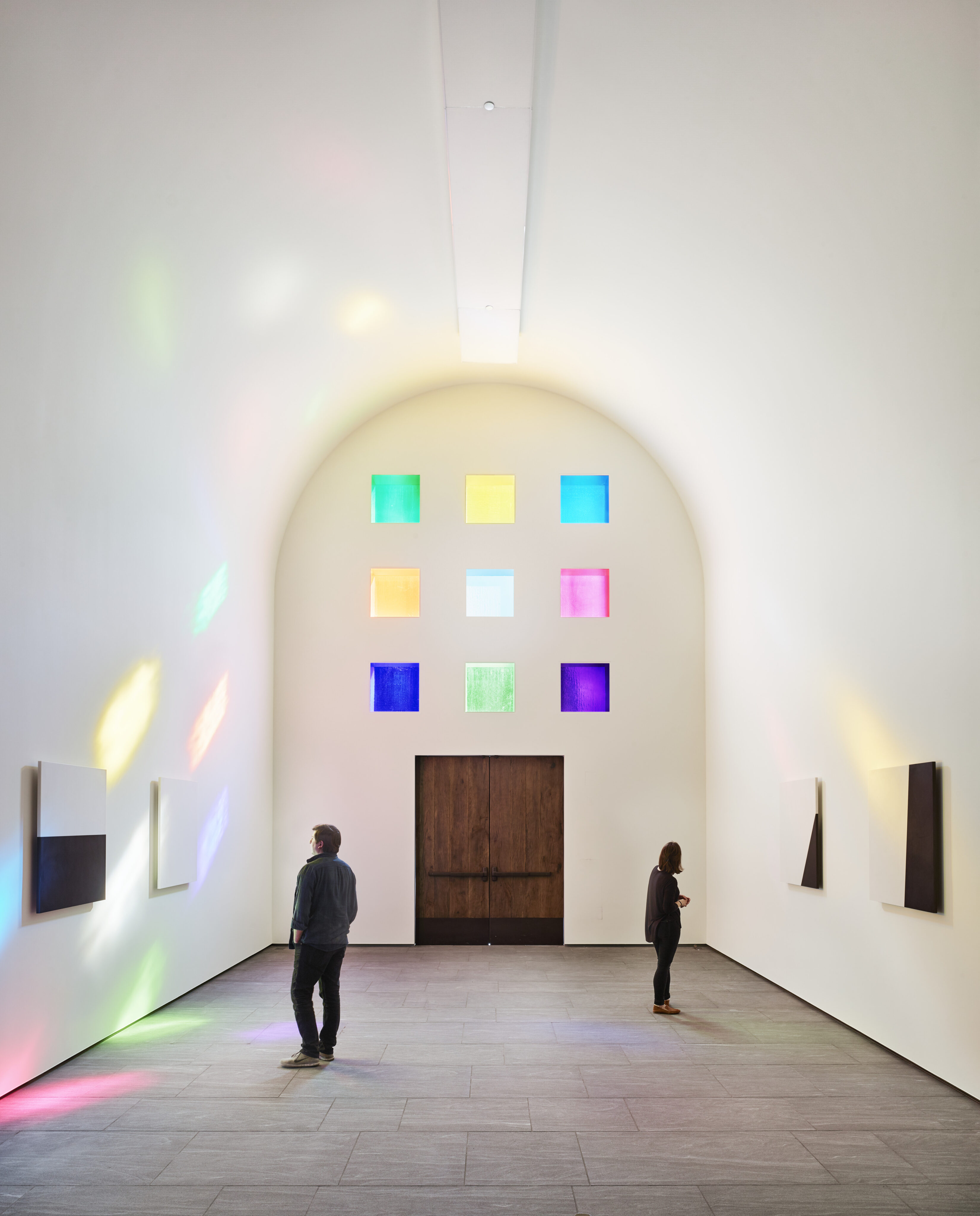 Two individuals stand in a minimalist gallery with colorful translucent squares on the back wall and abstract artwork on the side walls. Sunlight filters through the squares, casting colorful reflections reminiscent of Ellsworth Kelly’s Austin.