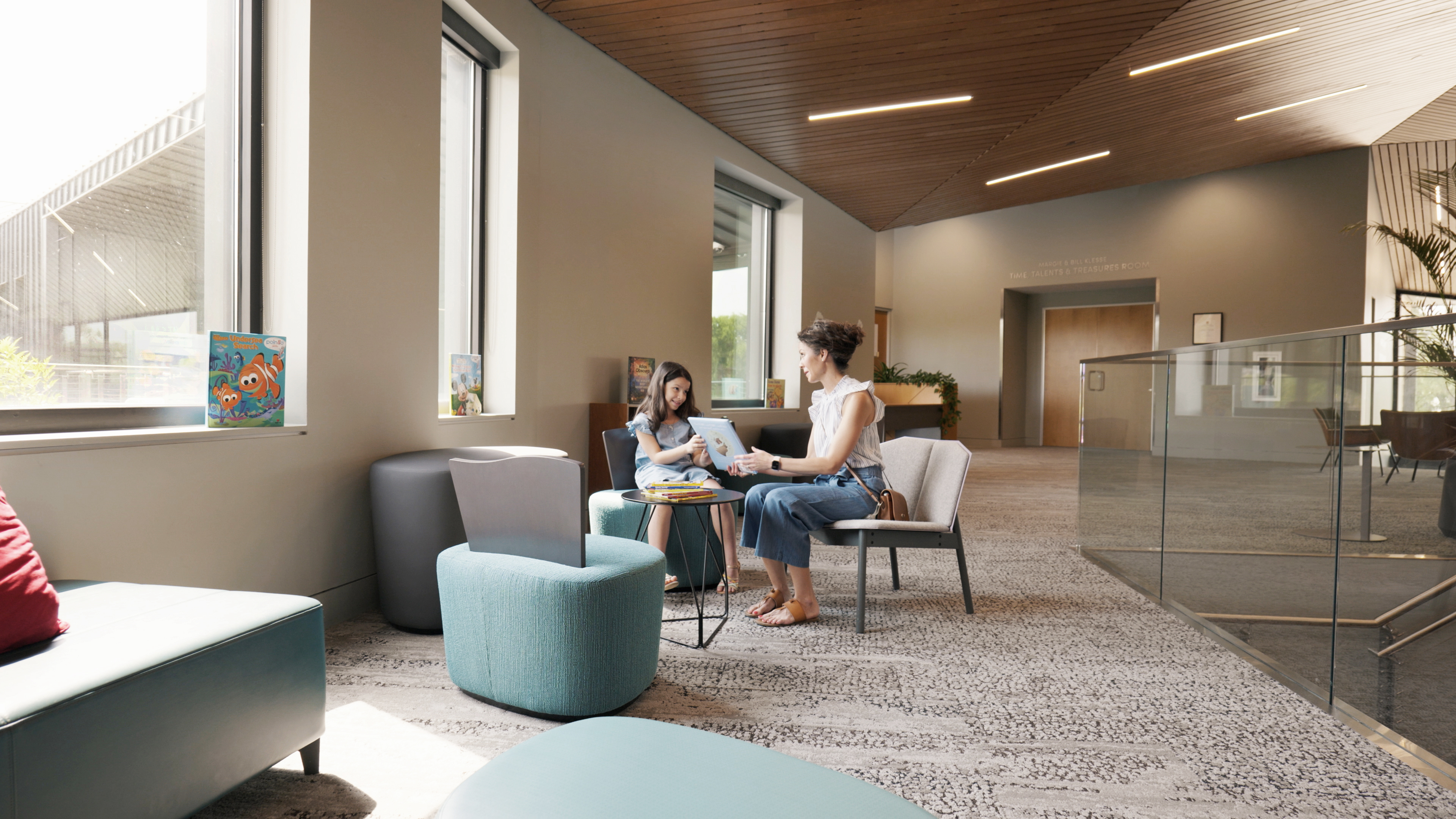 Two women are sitting and conversing in a modern, well-lit lounge area with large windows, contemporary furniture, and a high ceiling that creates an impactful atmosphere.