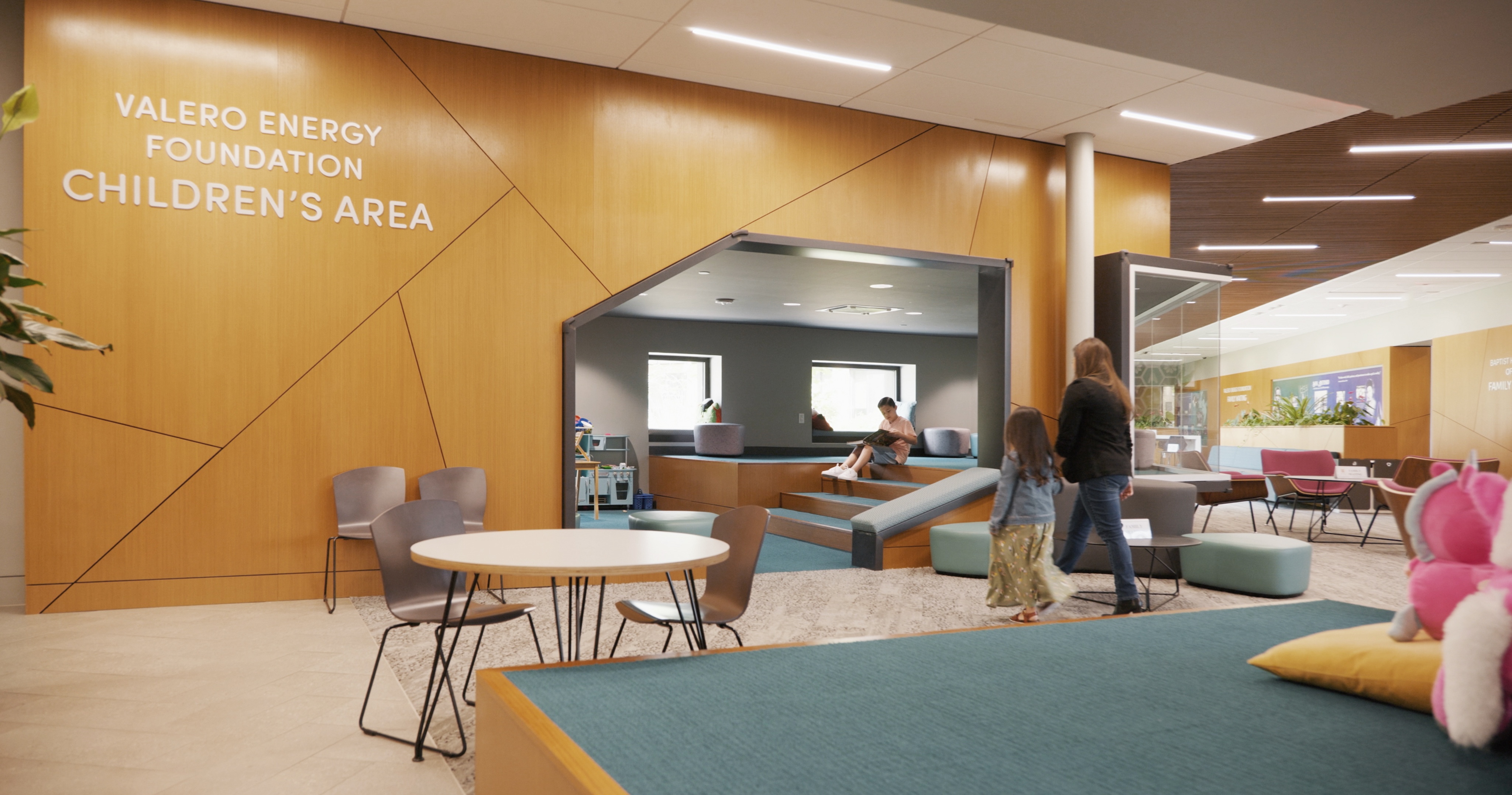 A modern children's area with seating, tables, and a play zone. A woman and a child walk towards the play area, and another child is seen playing on a slide. The wall reads "Valero Energy Foundation Children's Area" at the Harvey E. Najim Children & Family Center.