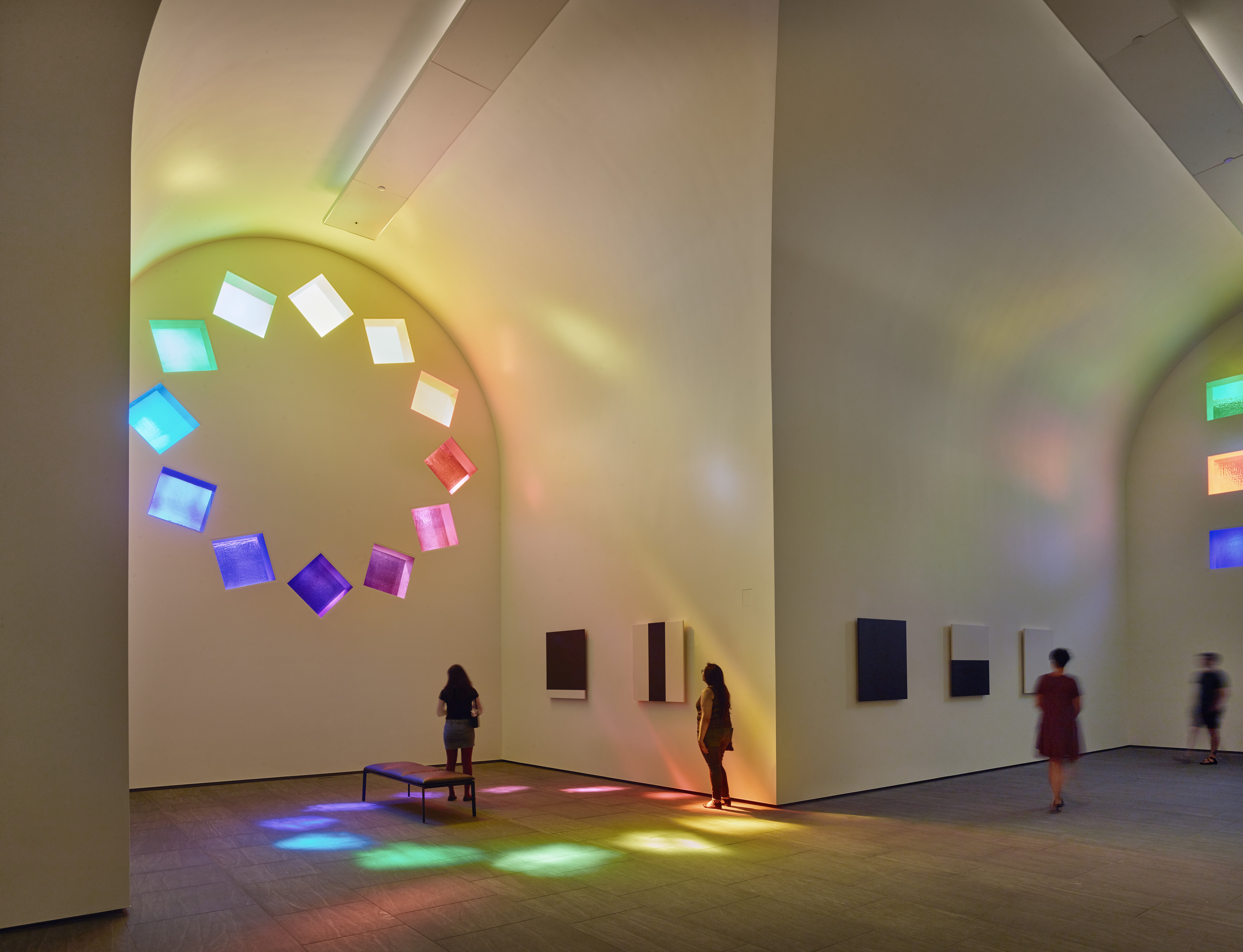 People observe the impactful, colorful, square-shaped light projections on the curved wall of an art gallery with high ceilings and minimalist décor.