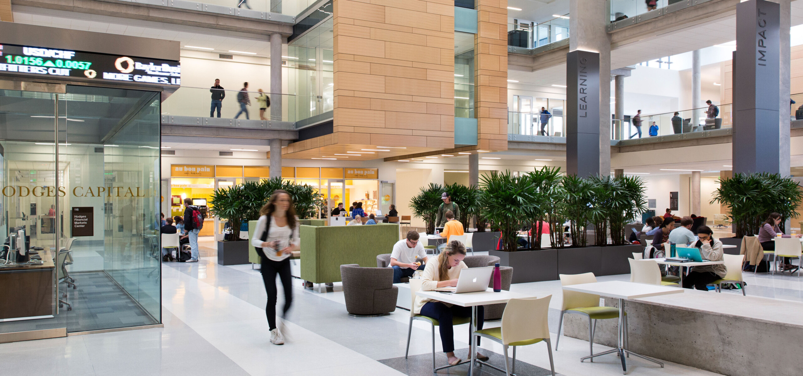 People are sitting and walking in the common area of a modern building with glass partitions, large plants, and multiple levels. Some are using laptops while others are engaged in conversation.