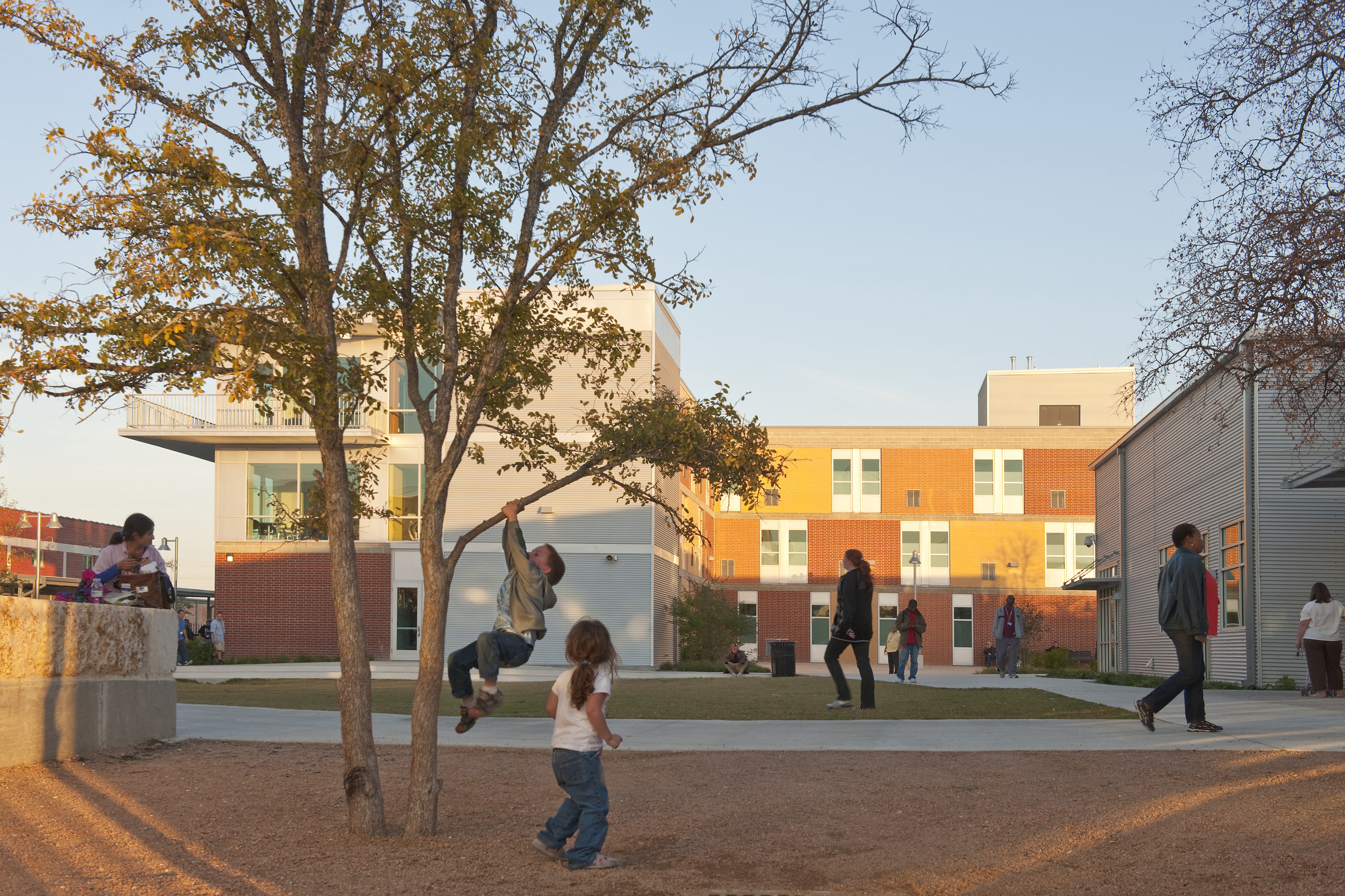 Children play near a tree while adults walk on the concrete path in a modern school courtyard with brick and white buildings in the background, creating a true Haven for Hope.