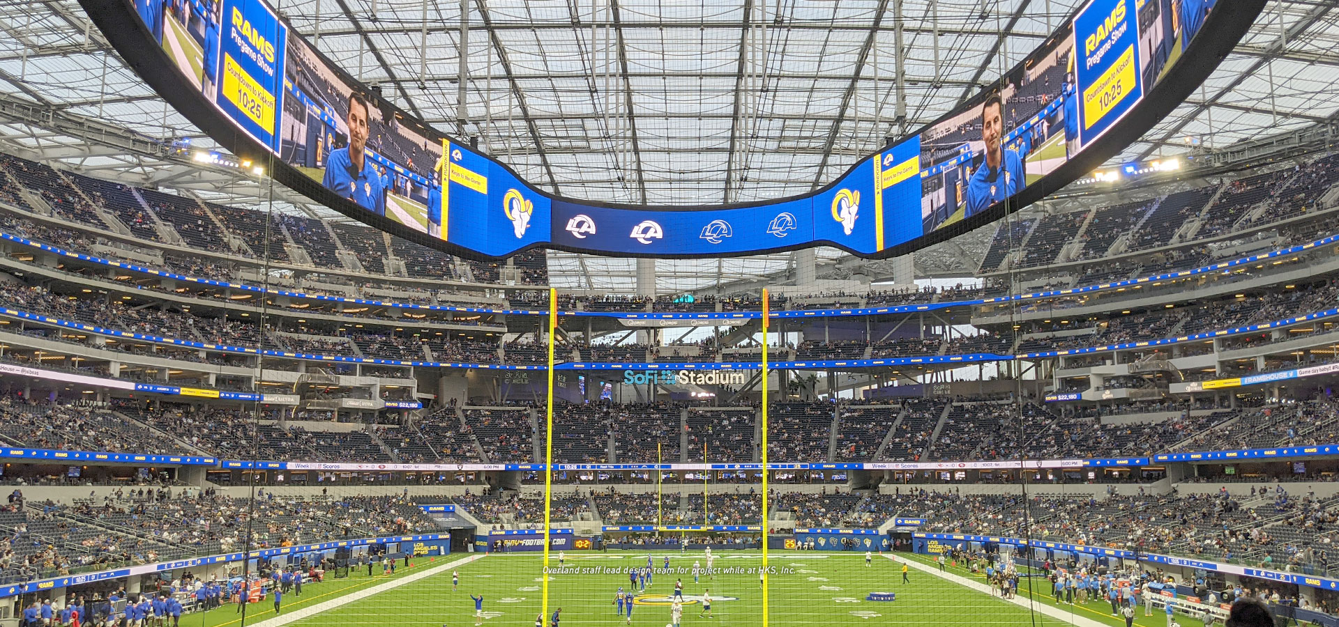 A football stadium filled with fans, with a large digital scoreboard and the field lined with players and coaches. The transparent roof, a marvel of modern engineering projects, provides ample natural light.