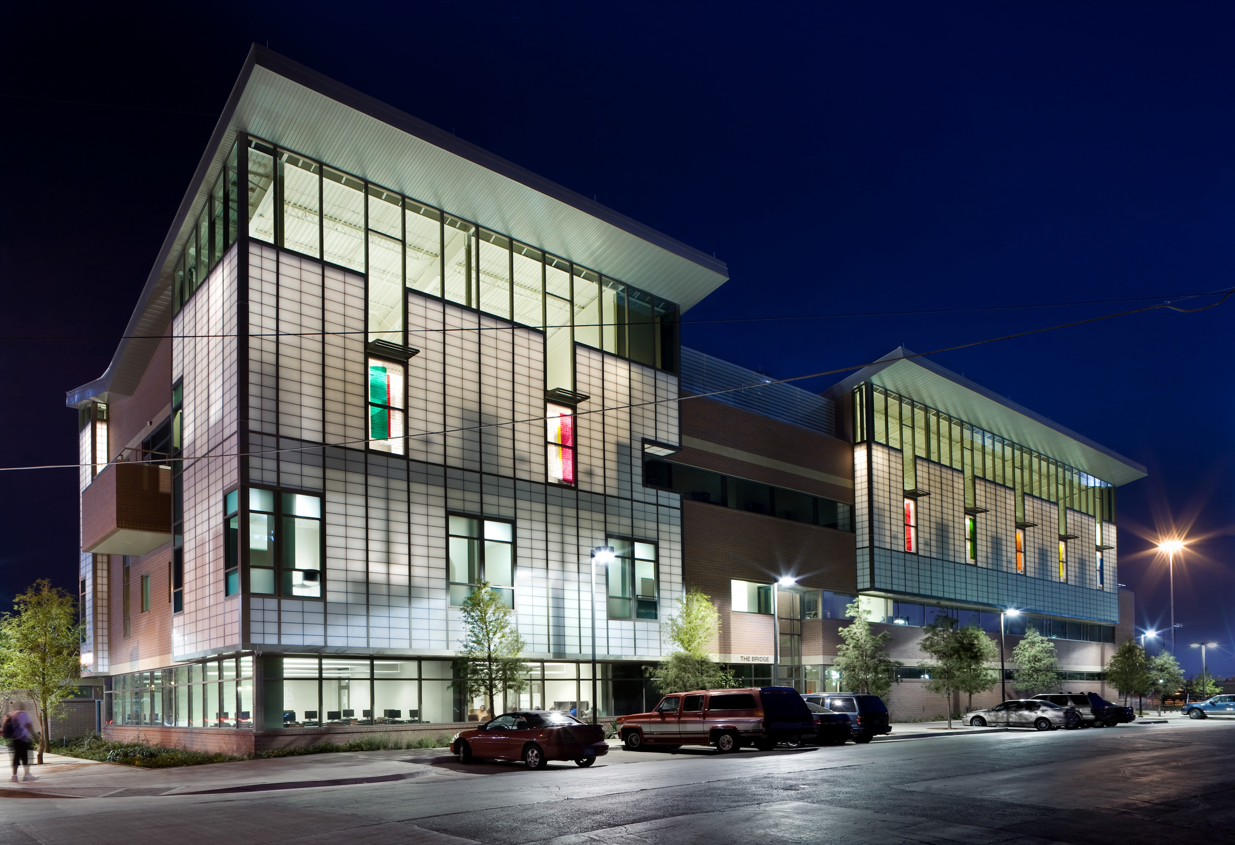 A modern, multi-story building illuminated at night, featuring large windows with colorful accents reminiscent of The Bridge. A few cars are parked in front and a person is walking nearby.