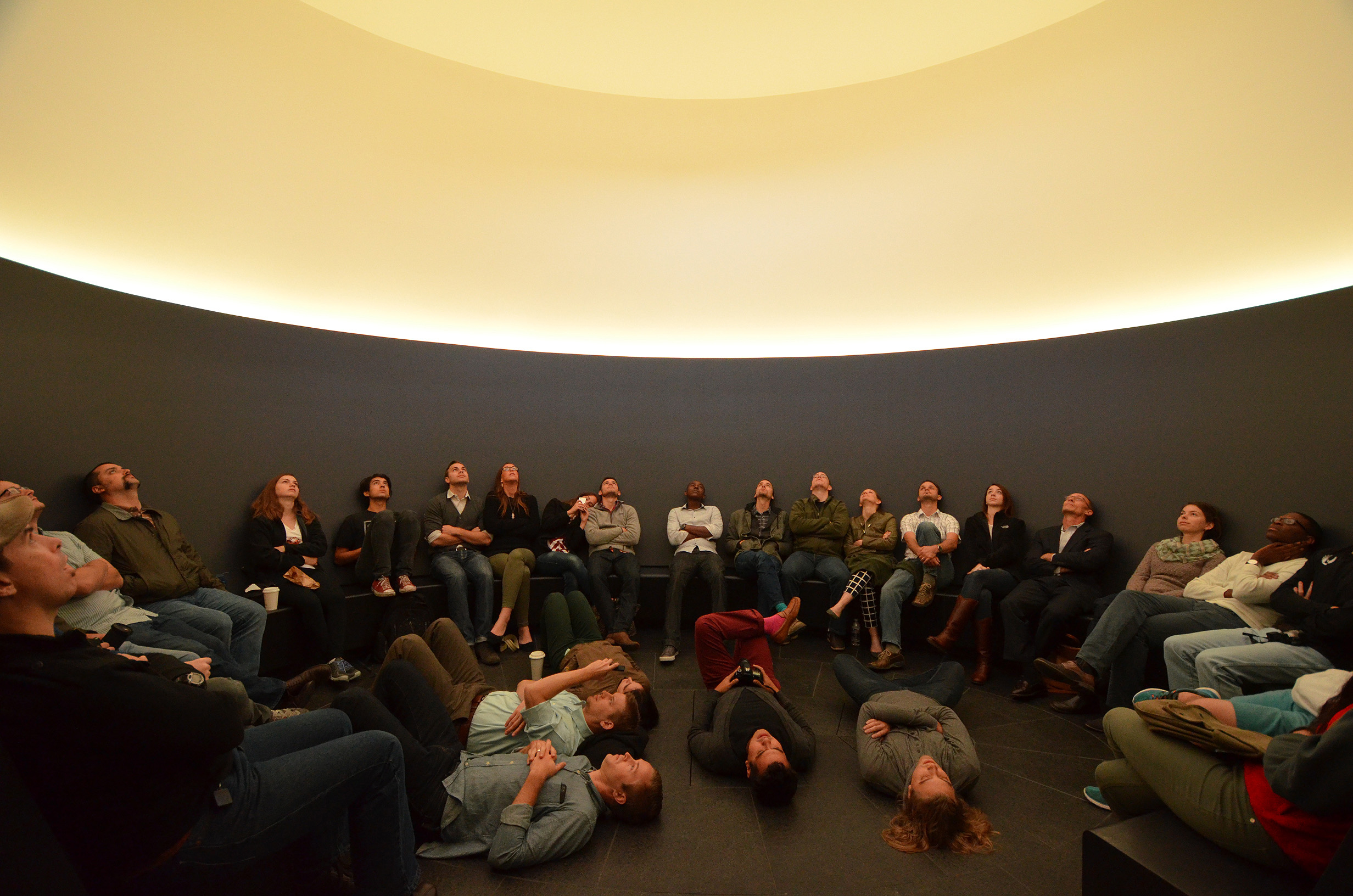 A group of people are sitting or lying on the floor in a dimly lit room, looking up at a circular, illuminated ceiling, feeling the profound impact of its mesmerizing glow.